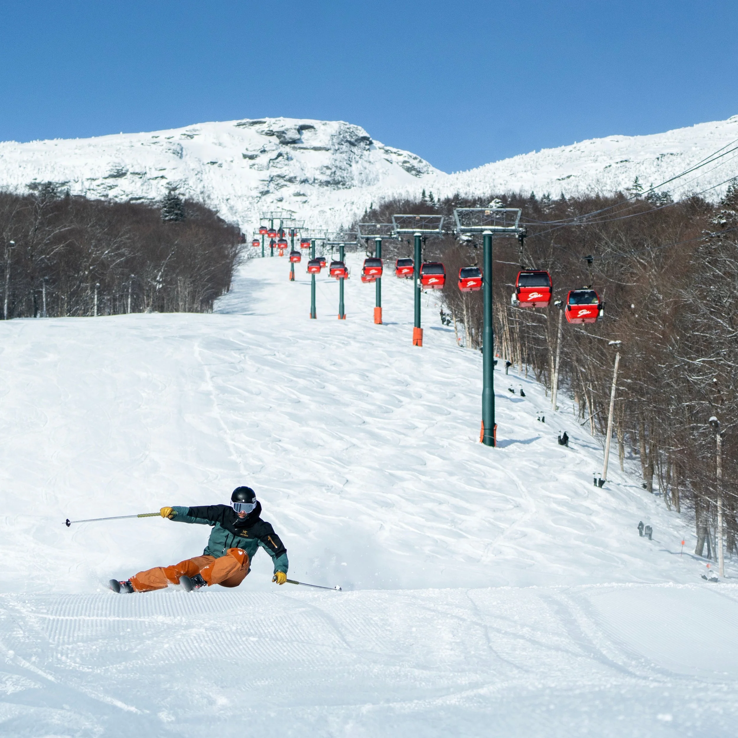 Two skiers falling on a snow-covered ski slope at a mountain resort with ski lifts, forest, and snow-capped peaks in the background.