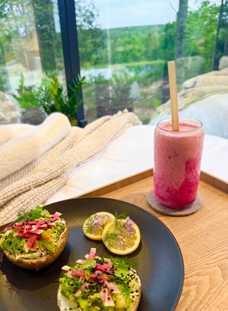 A breakfast tray with two bagel halves topped with avocado, herbs, and pickled vegetables, alongside three lemon slices garnished with herbs. There is a pink smoothie in a glass with a straw, placed on a wooden tray. The background shows a large wind