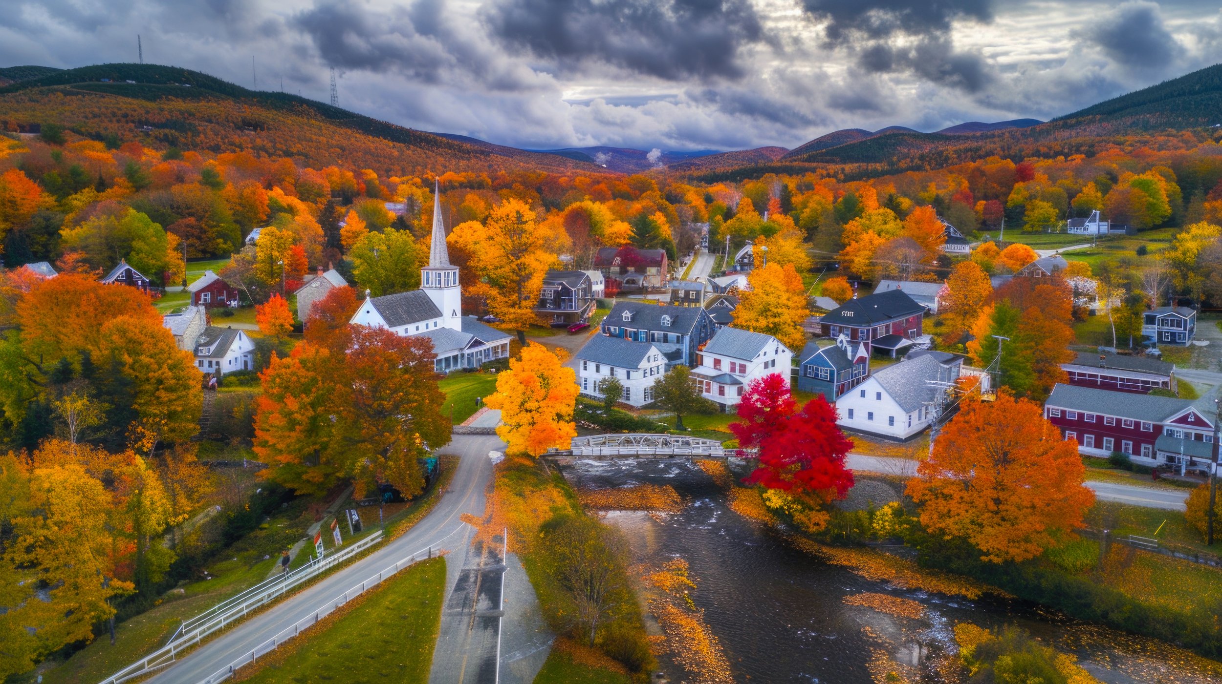 Aerial view of a small town in the fall with colorful trees, houses, a church, and mountains in the background.