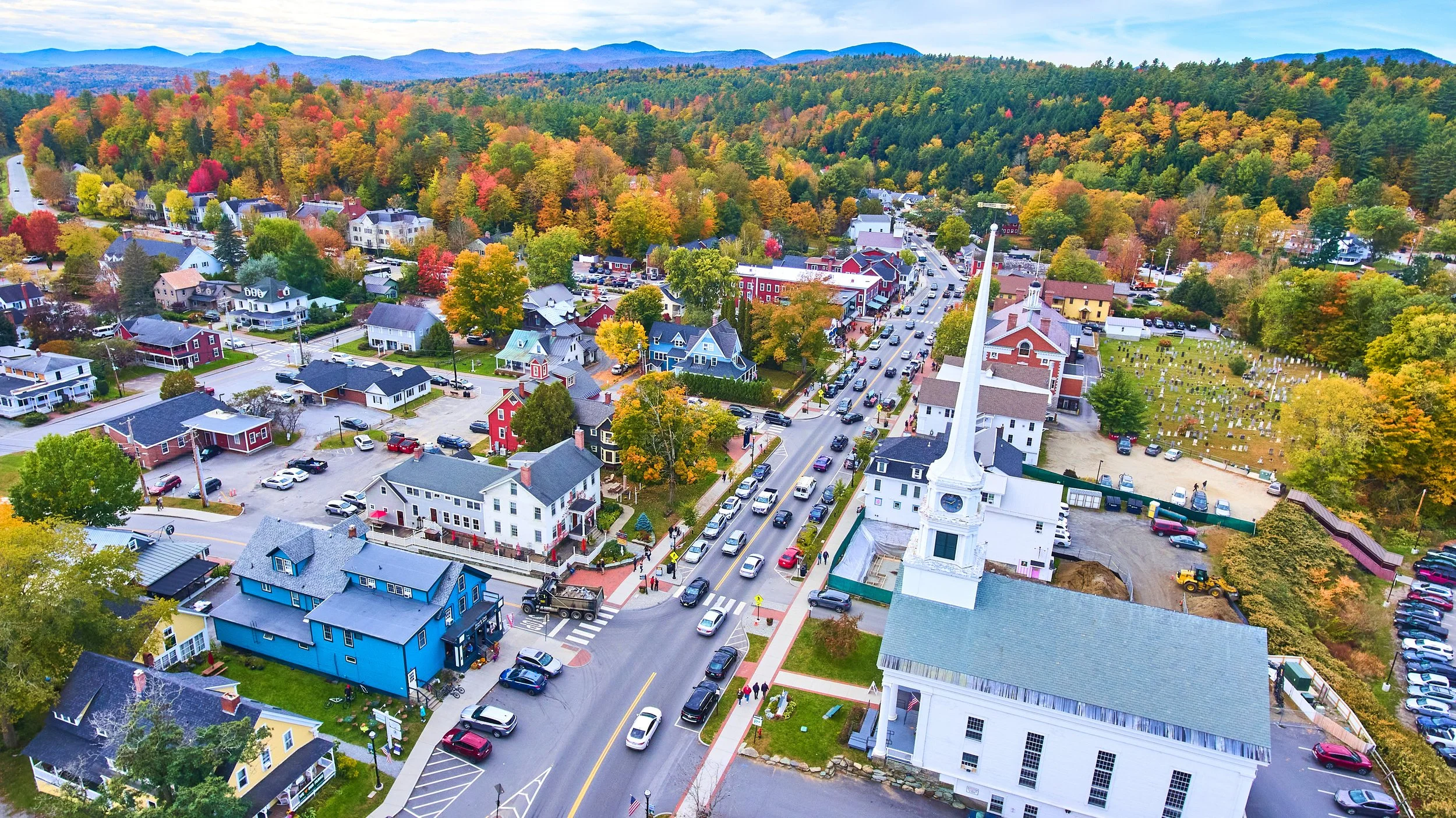 An aerial view of a small town with colorful houses, a church with a tall white steeple, and a tree-filled hillside in the background, during fall.