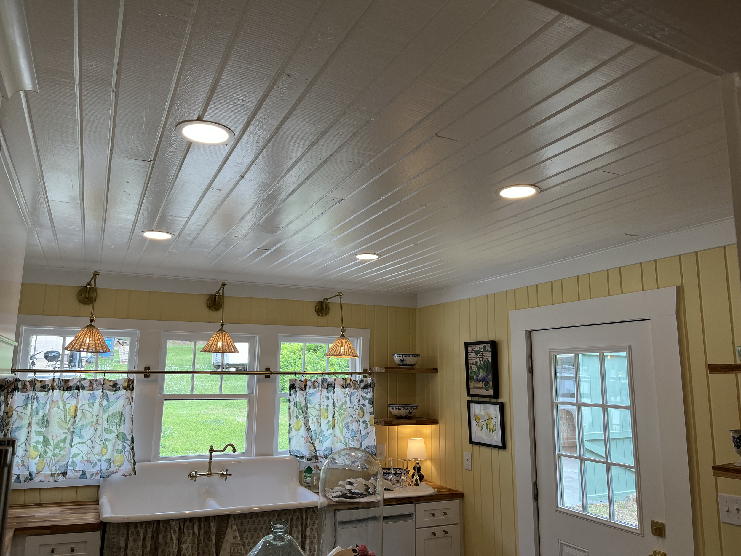 Living room with large window and yellow vertical paneling, farmhouse style chandelier, and kitchen area with a view of green lawn outside.