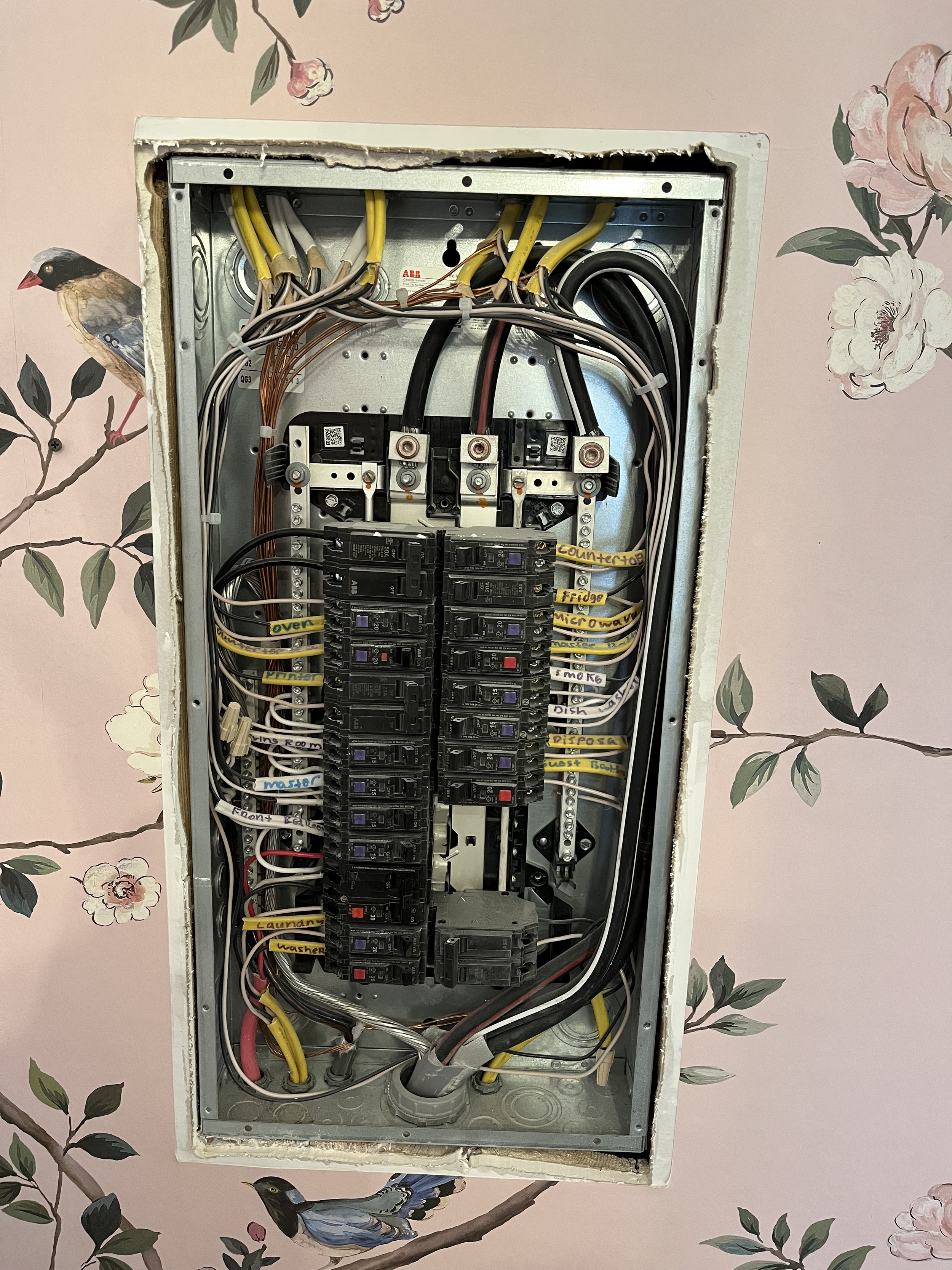 An open electrical panel with wires and circuit breakers, mounted on a wall decorated with pink floral wallpaper and bird illustrations.