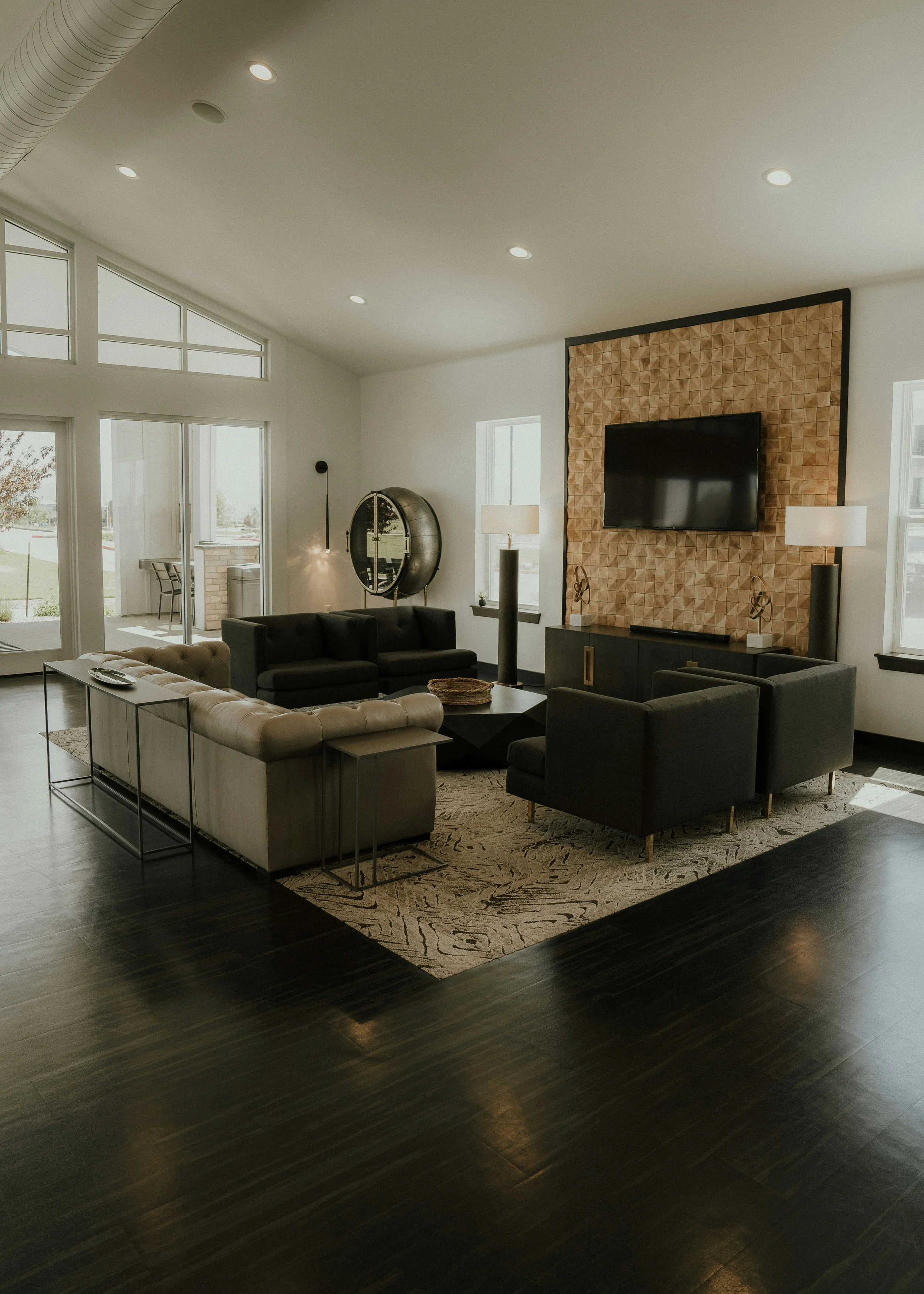 A modern living room with white walls, a large flat-screen TV mounted on a textured wooden panel, black sofas, glass side tables, and tall windows letting in natural light. There's a beige patterned rug on dark hardwood floors, and decorative lamps and sculptures.