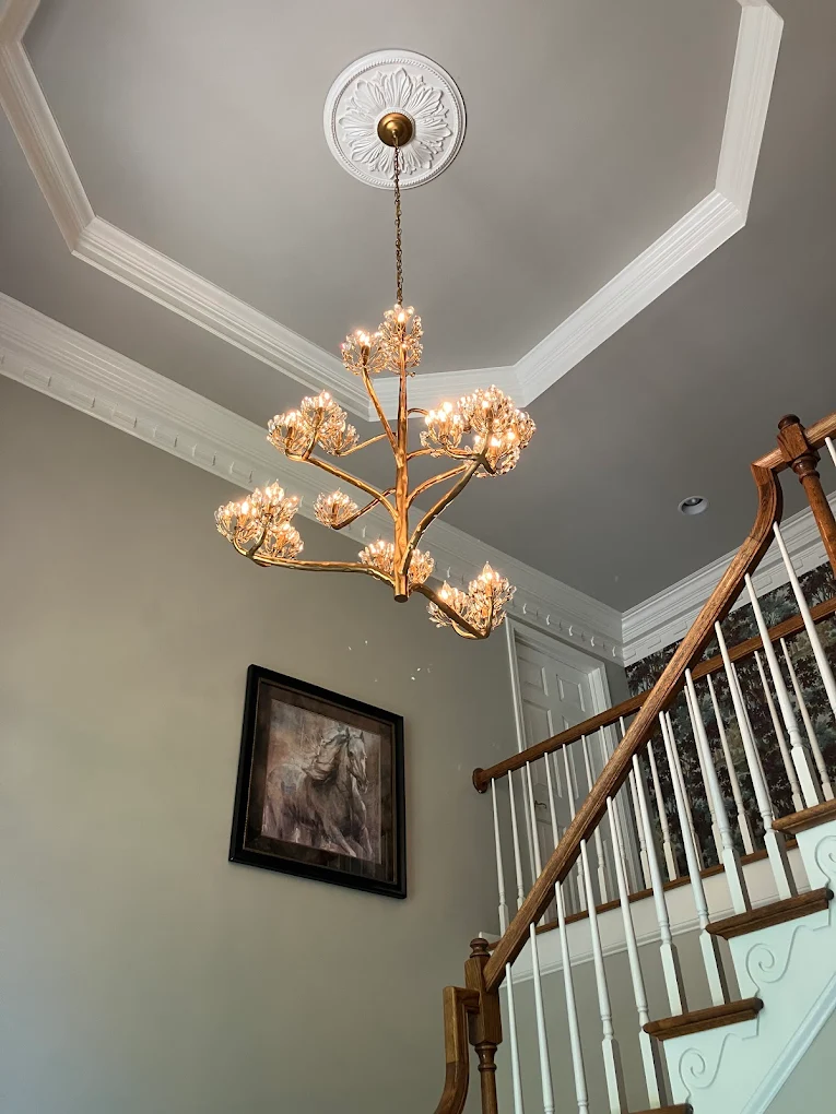 A chandelier with multiple branches and lights hangs from a decorative ceiling medallion in a foyer with a staircase. The space has crown molding and a framed picture of a horse on the wall.