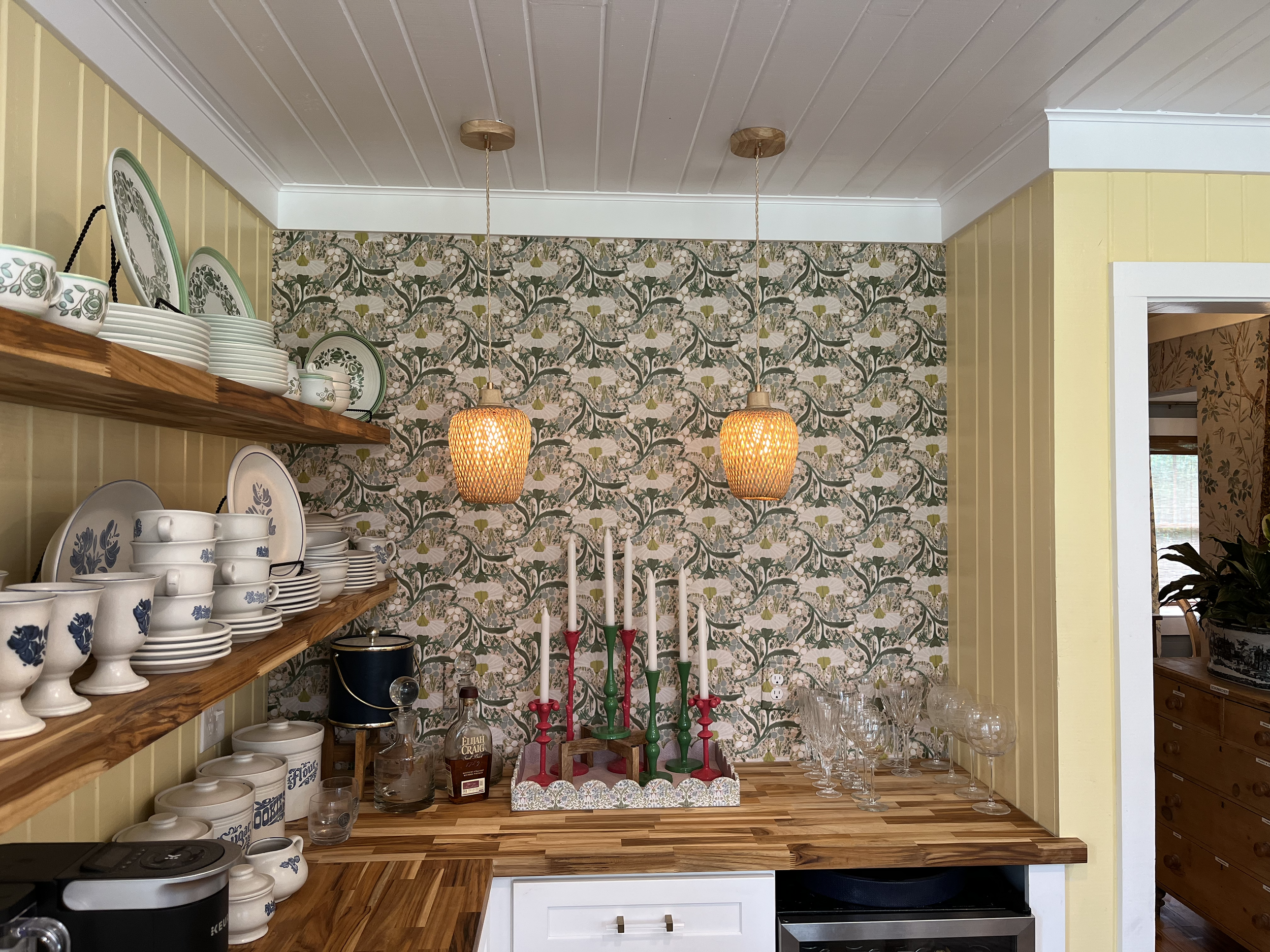 A kitchen corner with yellow beadboard walls, a wooden shelf filled with white and blue patterned dishware, green and pink candlesticks, glassware, and hanging woven pendant lights with a floral wallpaper background.