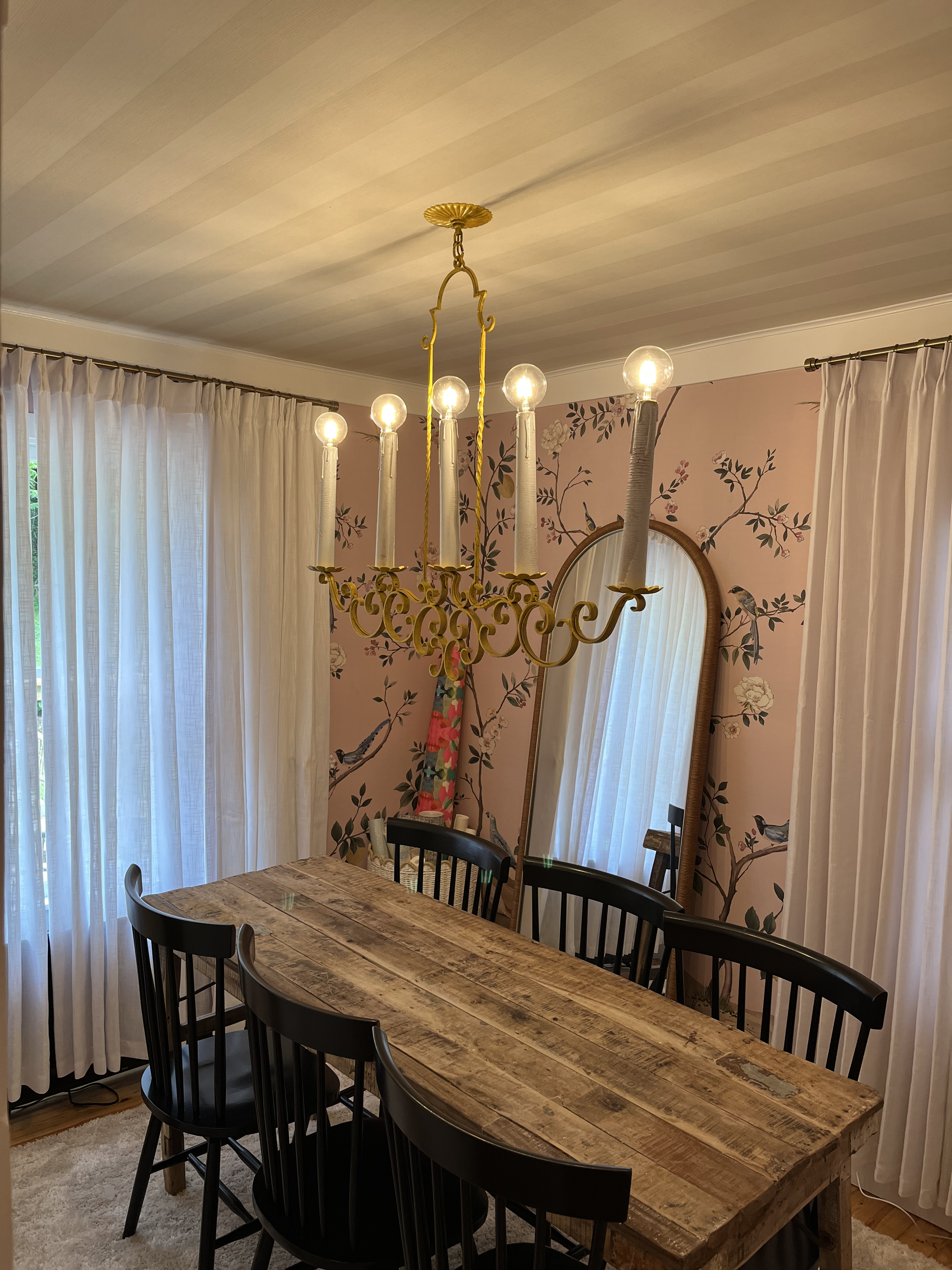A wooden dining table with black chairs in a room with pink wallpaper decorated with birds and flowers, white curtains, a mirror, and a chandelier with lit bulbs hanging from the ceiling.
