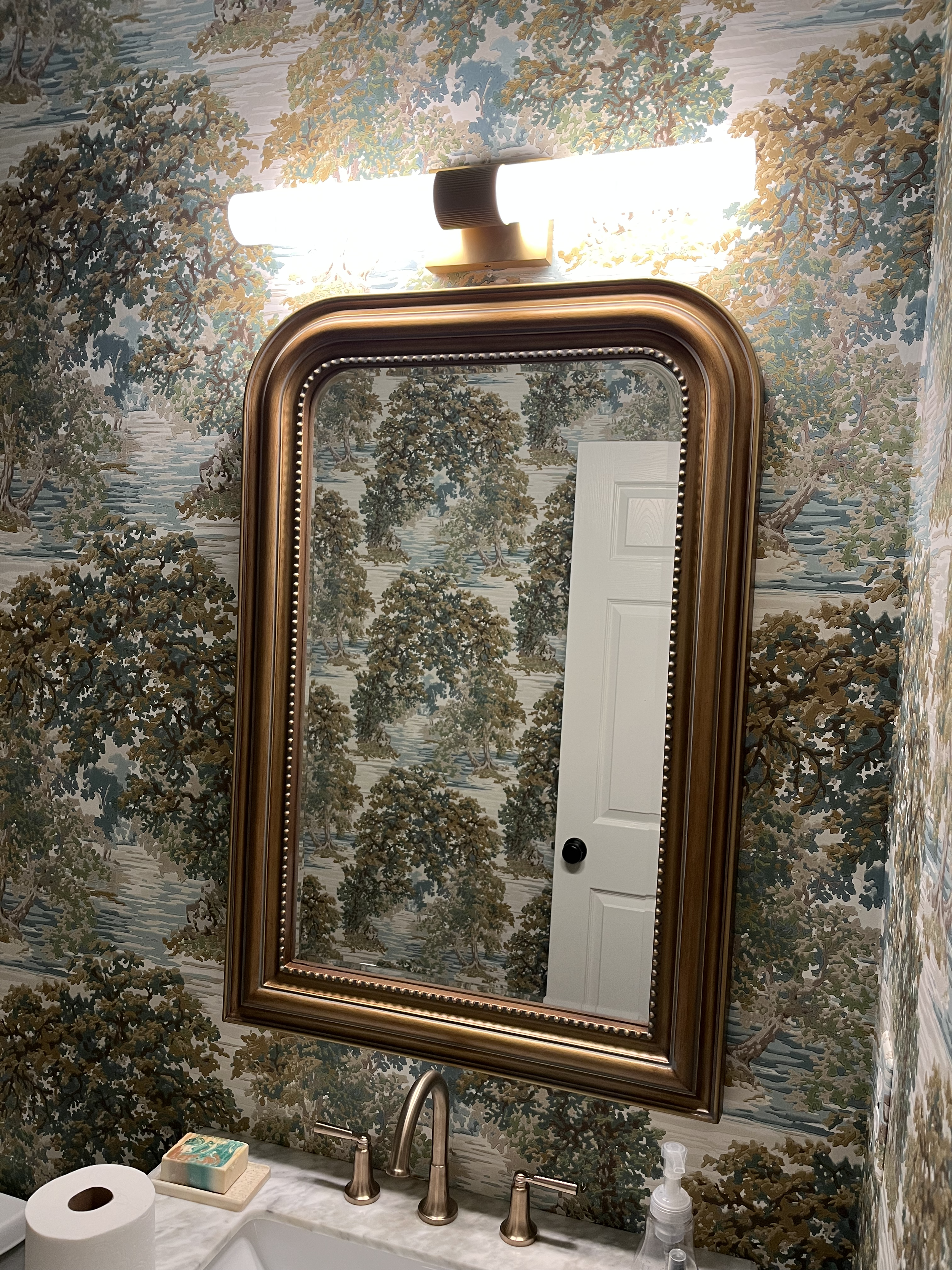 A bathroom mirror with a wooden frame, mounted on a wall covered with woodland-themed wallpaper, above a sink with a marble countertop, soap, and hand sanitizer.
