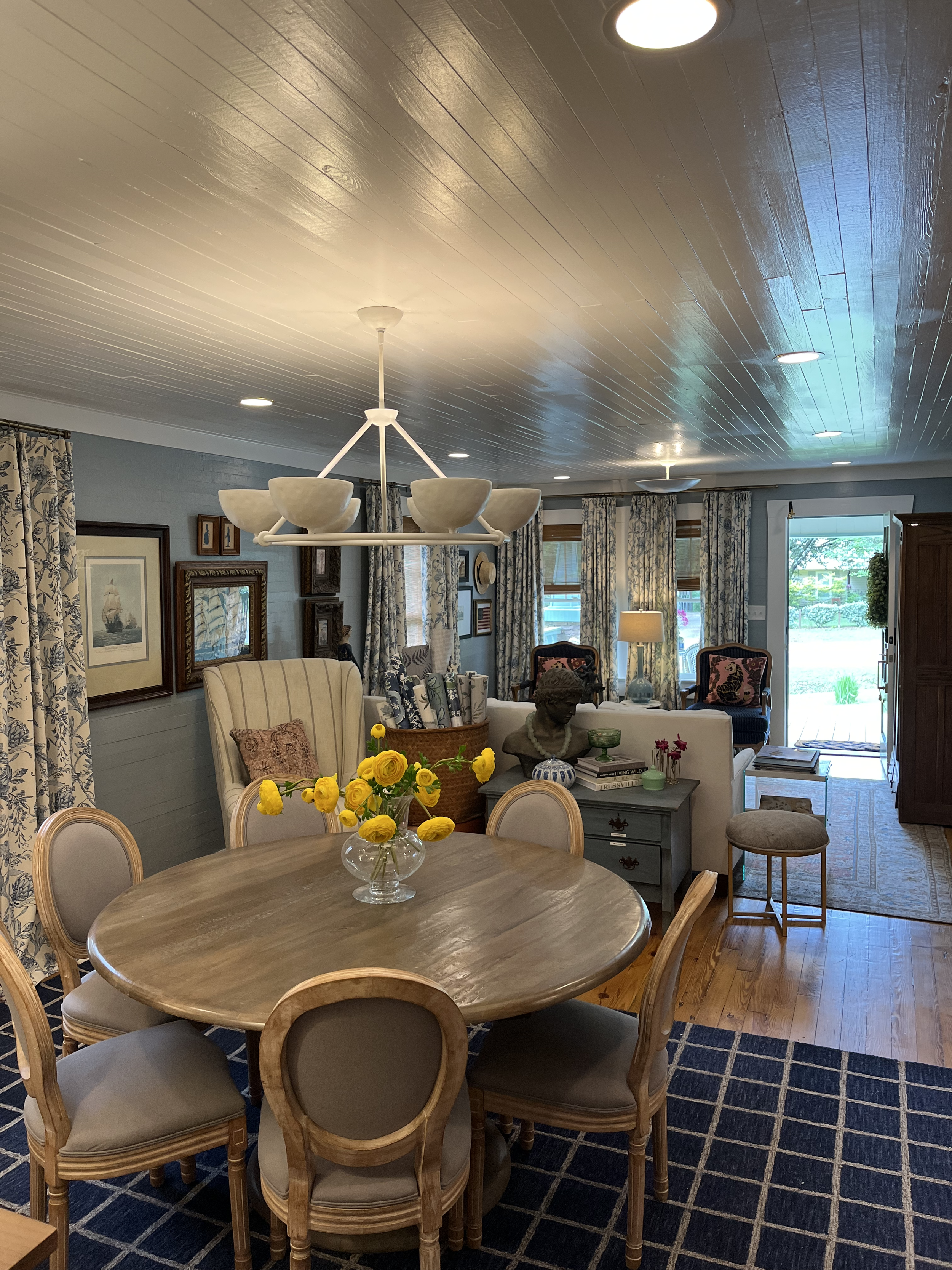 A dining room with a wooden table, surrounded by wooden chairs with beige and gray cushions. A vase with yellow flowers sits on the table. The background features a living area with chairs, a sofa, paintings on the wall, curtains, and windows letting