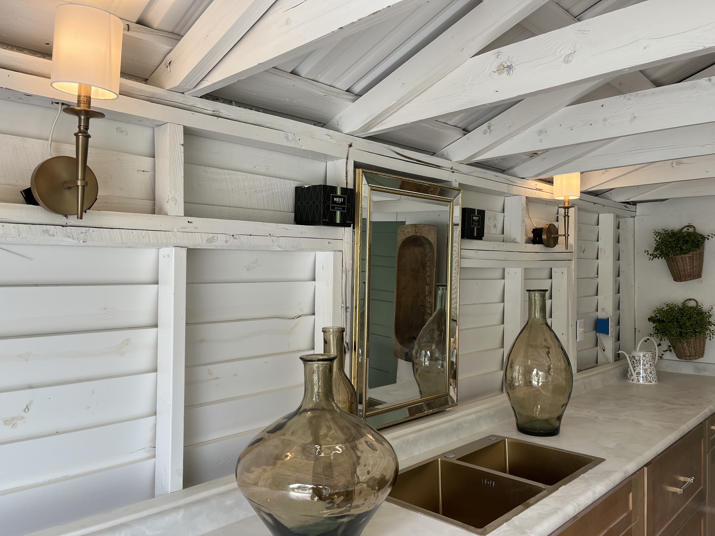 Interior of a room with white wooden walls and ceiling, featuring brass wall sconces with lampshades, decorative glass vases on a marble countertop, a gold-framed mirror, and potted plants.