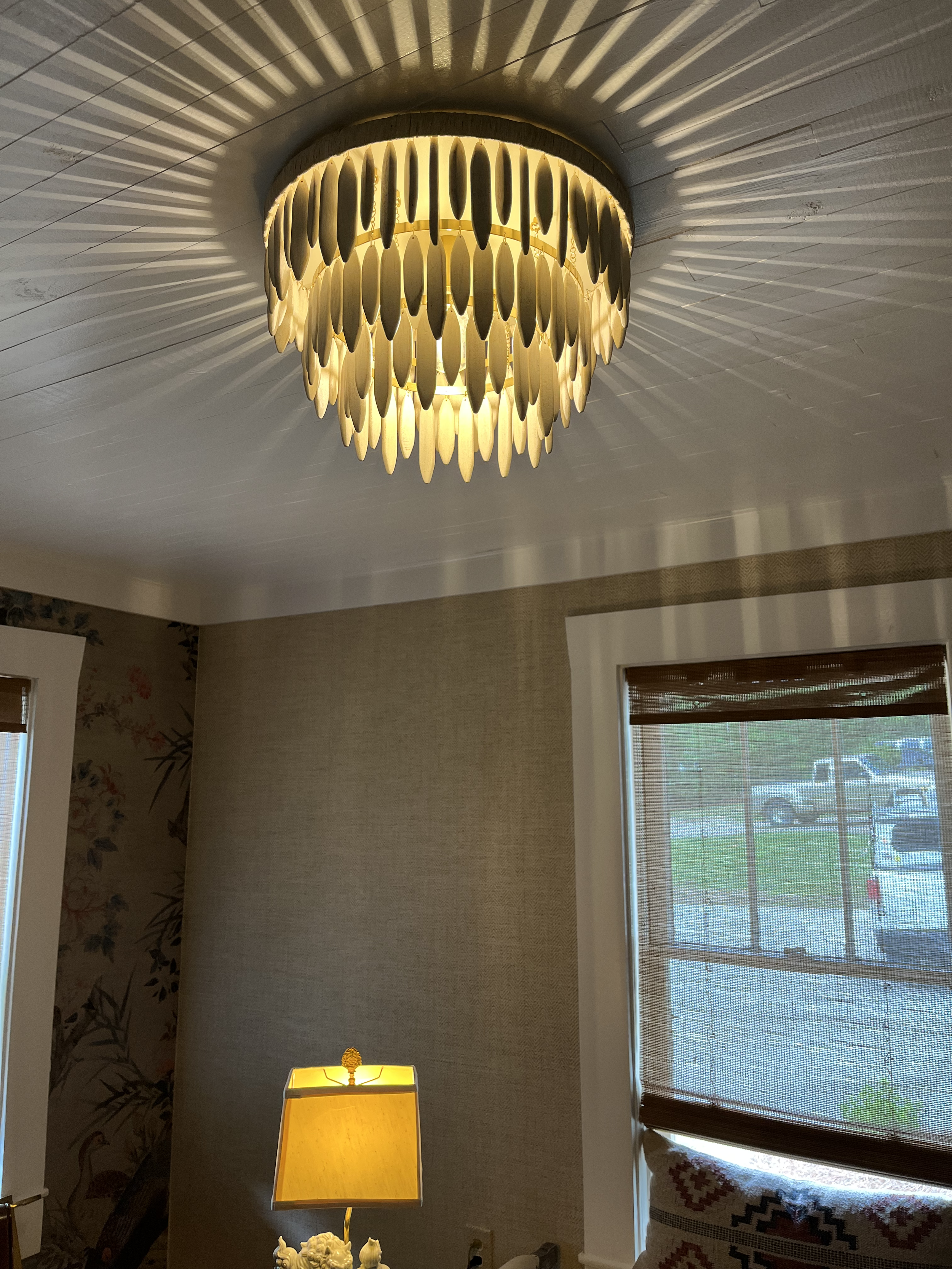 A ceiling light fixture with a gold frame and elongated oval decorations hanging downward, creating a shadow pattern on the ceiling. A table lamp with a yellow shade is visible below, along with part of a window with blinds and a woven curtain on the