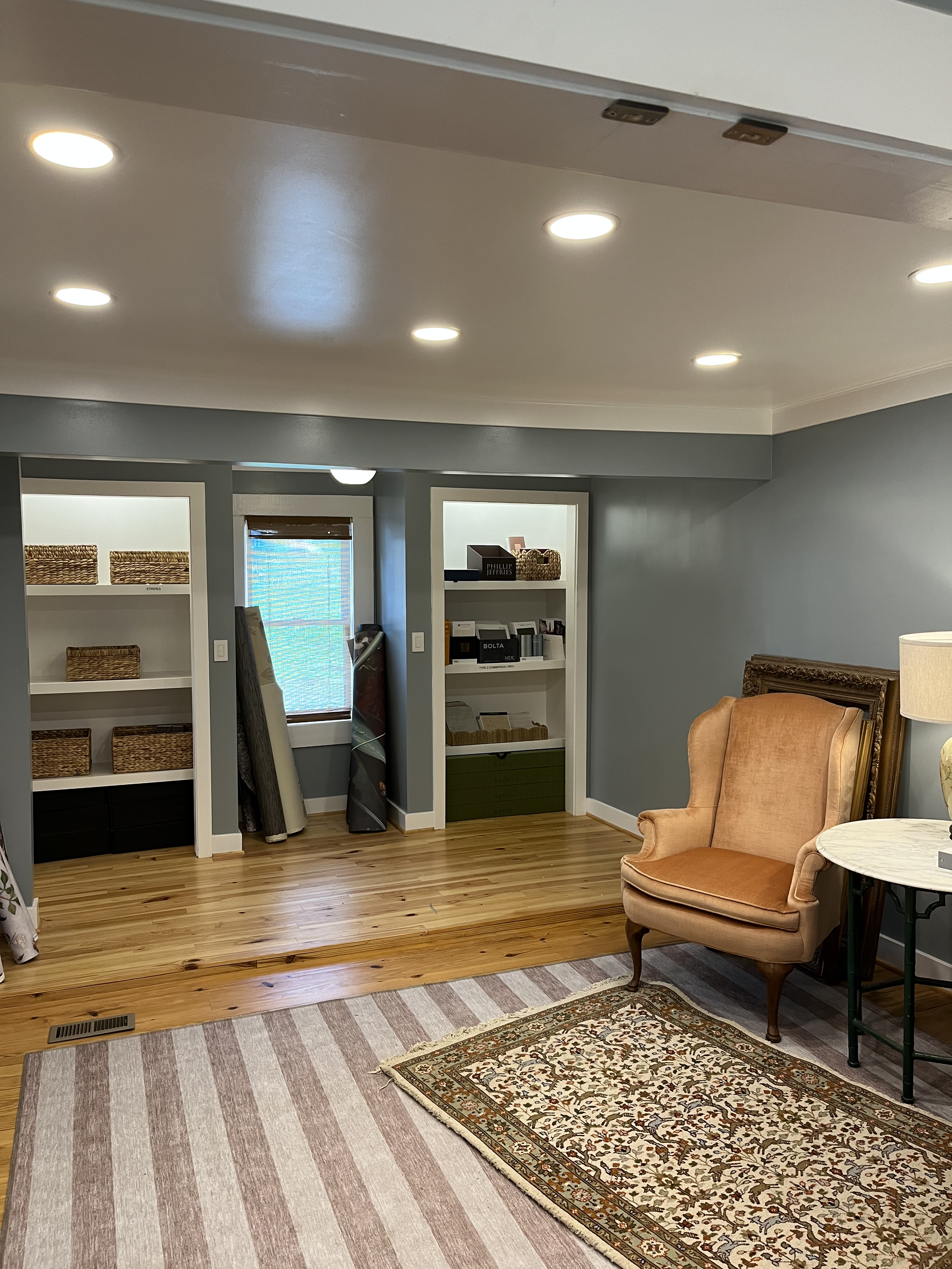 Living room with hardwood floors, beige and gray walls, white bookshelves, a window with a brown blind, a pink upholstered armchair, a side table with a lamp, and a decorative rug.