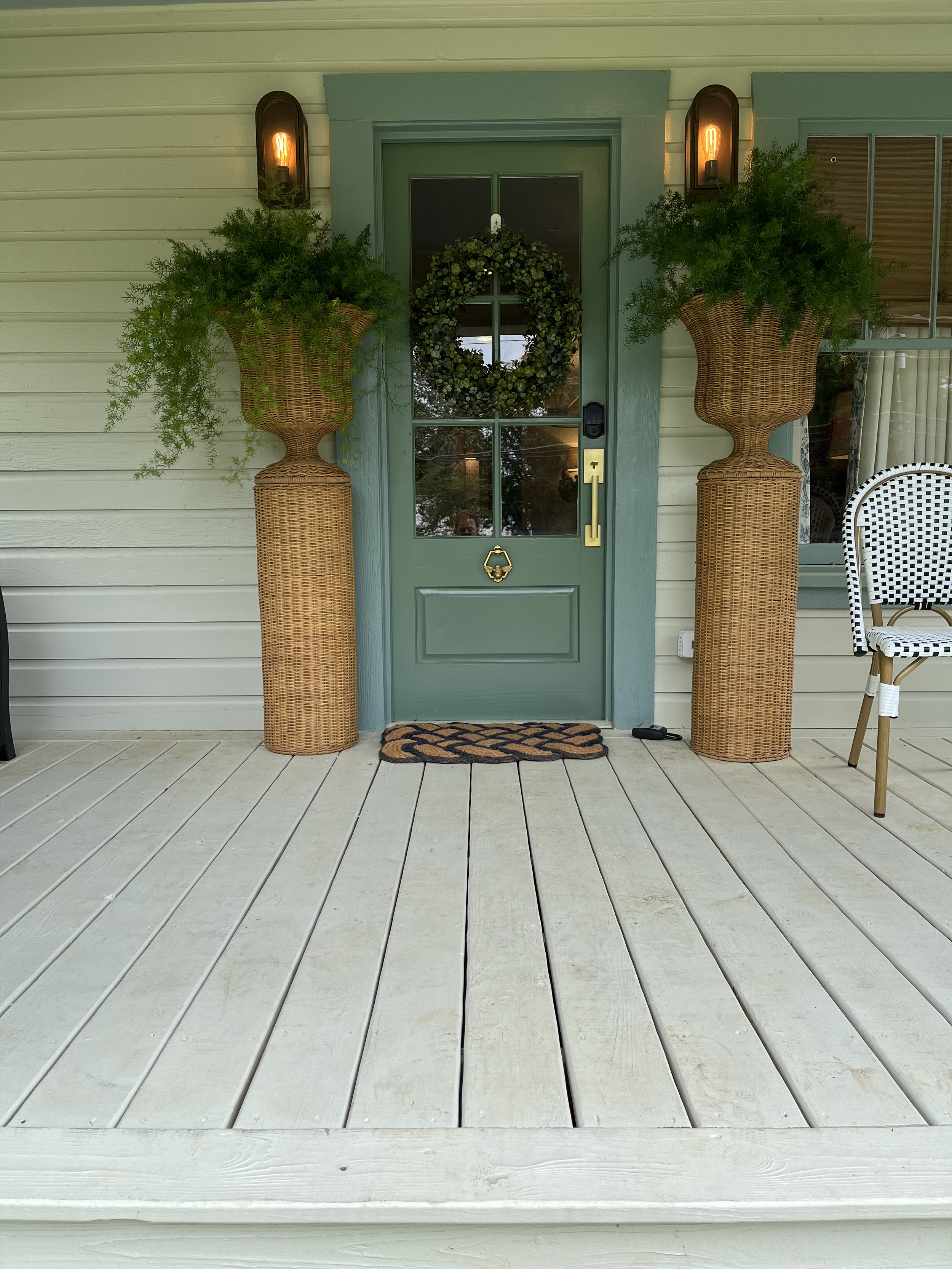 A front door with a green frame and glass panels, decorated with a wreath. Large wicker planters with greenery sit on either side of the door, and wall-mounted lights are above each planter. A striped chair is to the right, and a doormat is in front 
