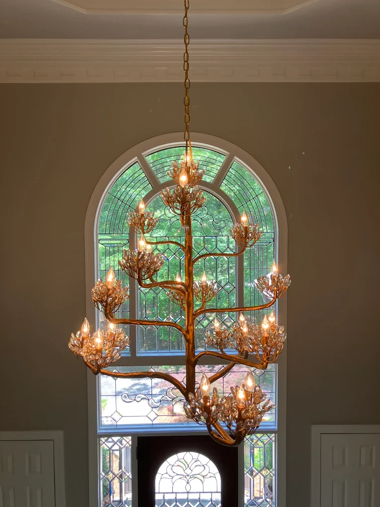 Large chandelier designed like a tree with multiple branches and crystal-like light fixtures hanging inside an elegant room with large arched window.