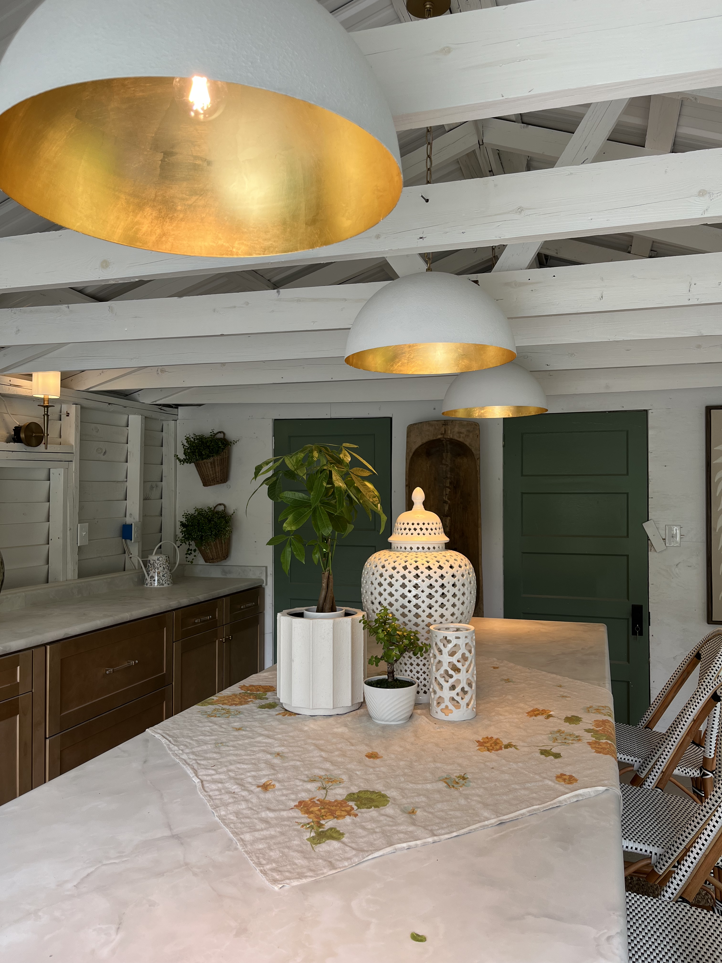 Inside a room with white painted wooden beams and ceiling, a dining table with a floral tablecloth, decorative vases and plants, and pendant lights hanging from the ceiling.