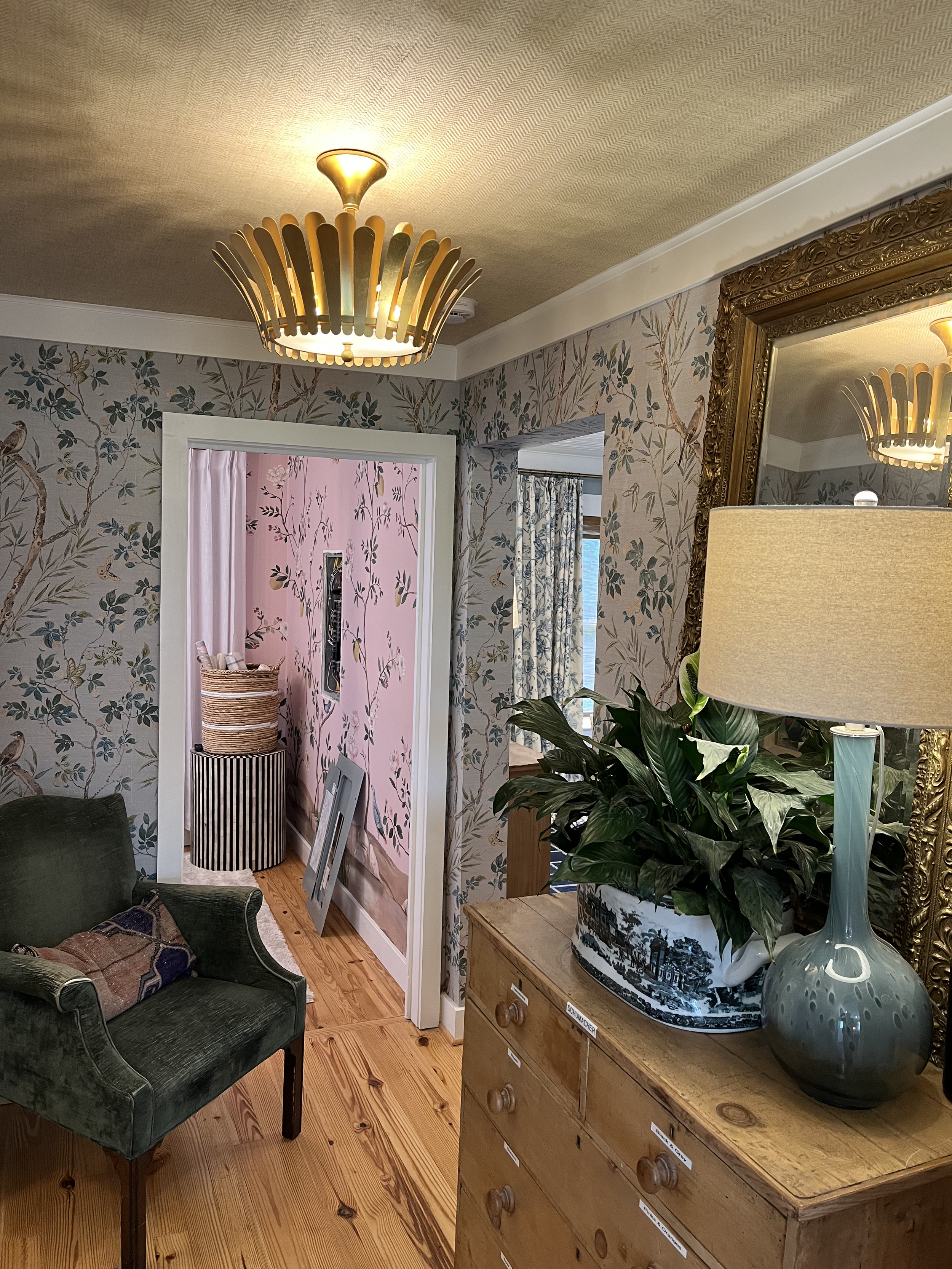 A cozy room with floral wallpaper, a large ornate mirror, a wooden dresser with plants and a lamp, a green armchair, and an open doorway with pink walls and a basket inside.