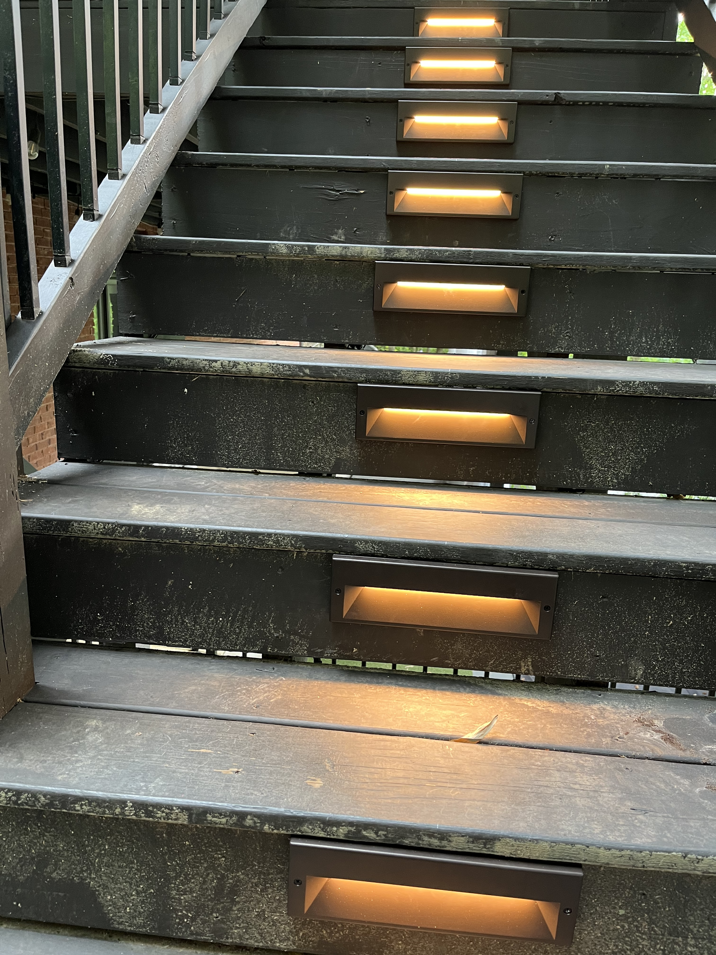 Close-up view of black outdoor stairs with built-in yellow lighting, showing some wear and dirt on the steps.