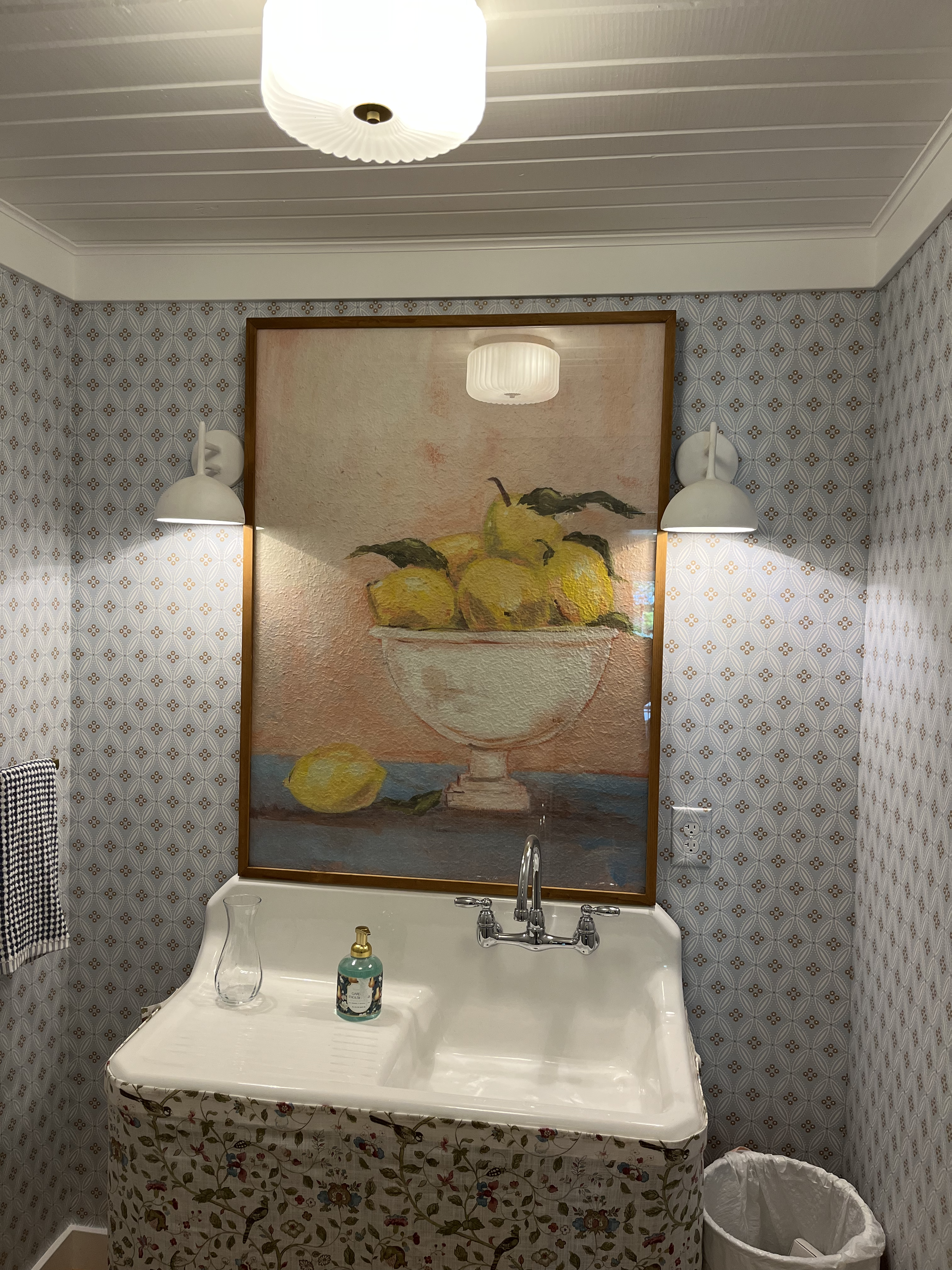 Bathroom with a vintage sink, floral curtain, patterned wallpaper, and a painting of lemons in a bowl on the wall. There are two white wall-mounted lamps, a bottle of hand soap, a clear glass, and a trash bin.