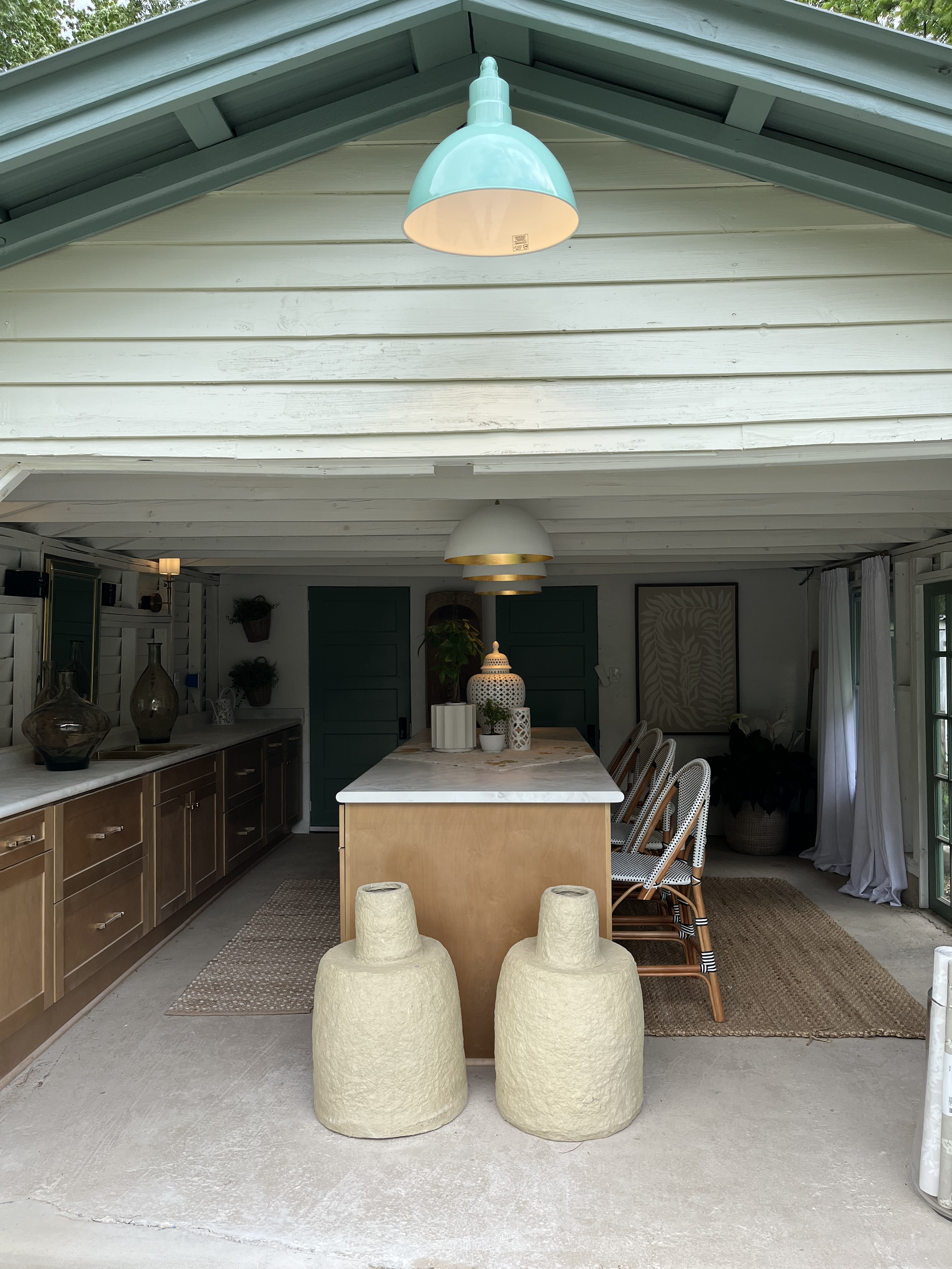 An outdoor covered patio with a ceiling light, a kitchen island, barstools, and decorative vases and plants on the counter.