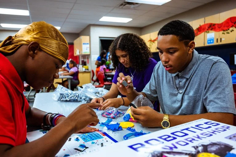 Group of kids building robots with plastic parts and tools at a robotics workshop in a community center.