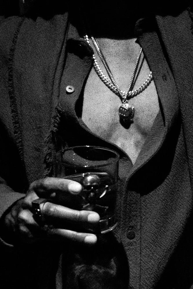 Persona usando collares en capas mientras sostiene un vaso con una bebida oscura. Viste una camisa parcialmente desabotonada y una chaqueta, en una fotografía en blanco y negro que transmite un estilo elegante y auténtico.