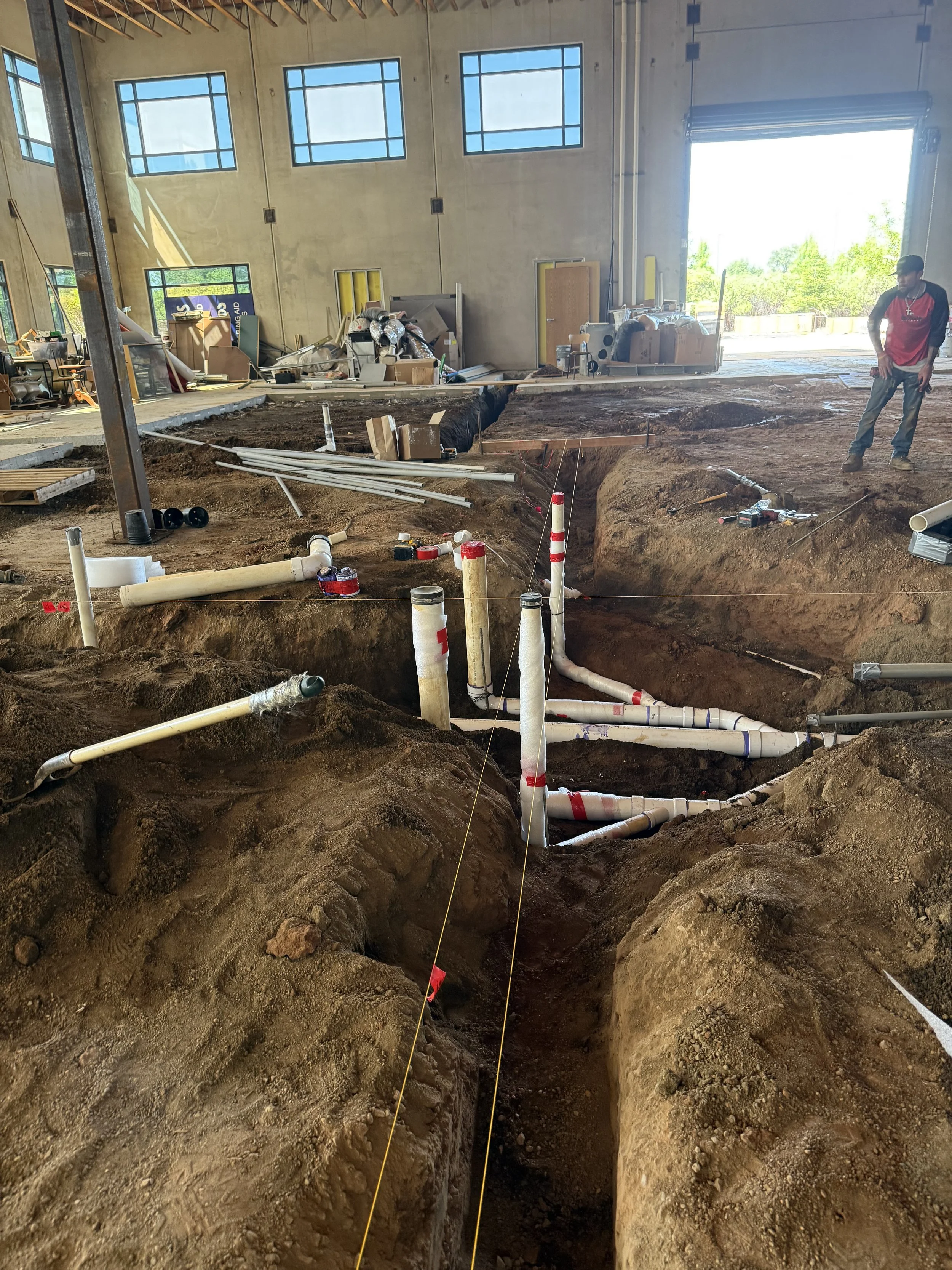 Construction site with exposed underground pipes and a worker standing nearby, inside a large partially constructed commercial building with high walls and multiple windows.