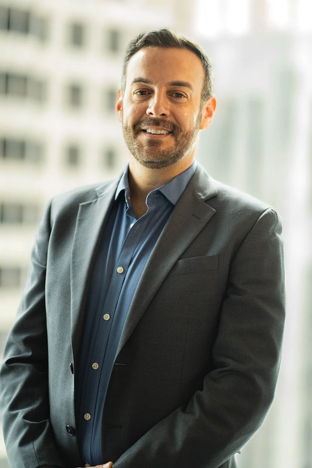 A man in a gray suit jacket and blue dress shirt smiling in an office building with windows in the background.