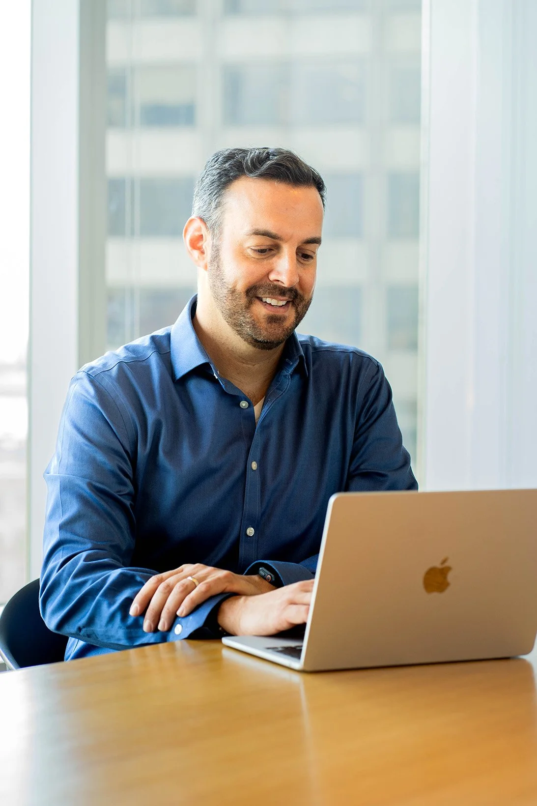 Man in blue shirt working on a silver Apple MacBook at a wooden table in a bright office with large windows.