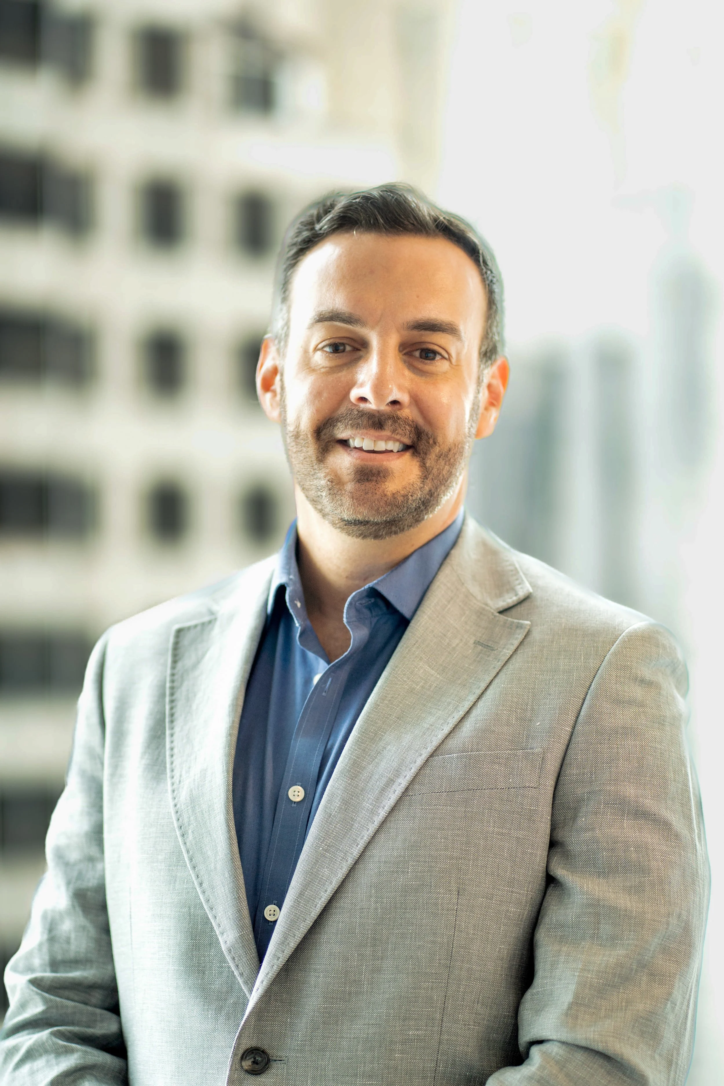 A man with a beard and dark hair, wearing a beige suit jacket over a blue shirt, smiling in front of a blurred cityscape background.