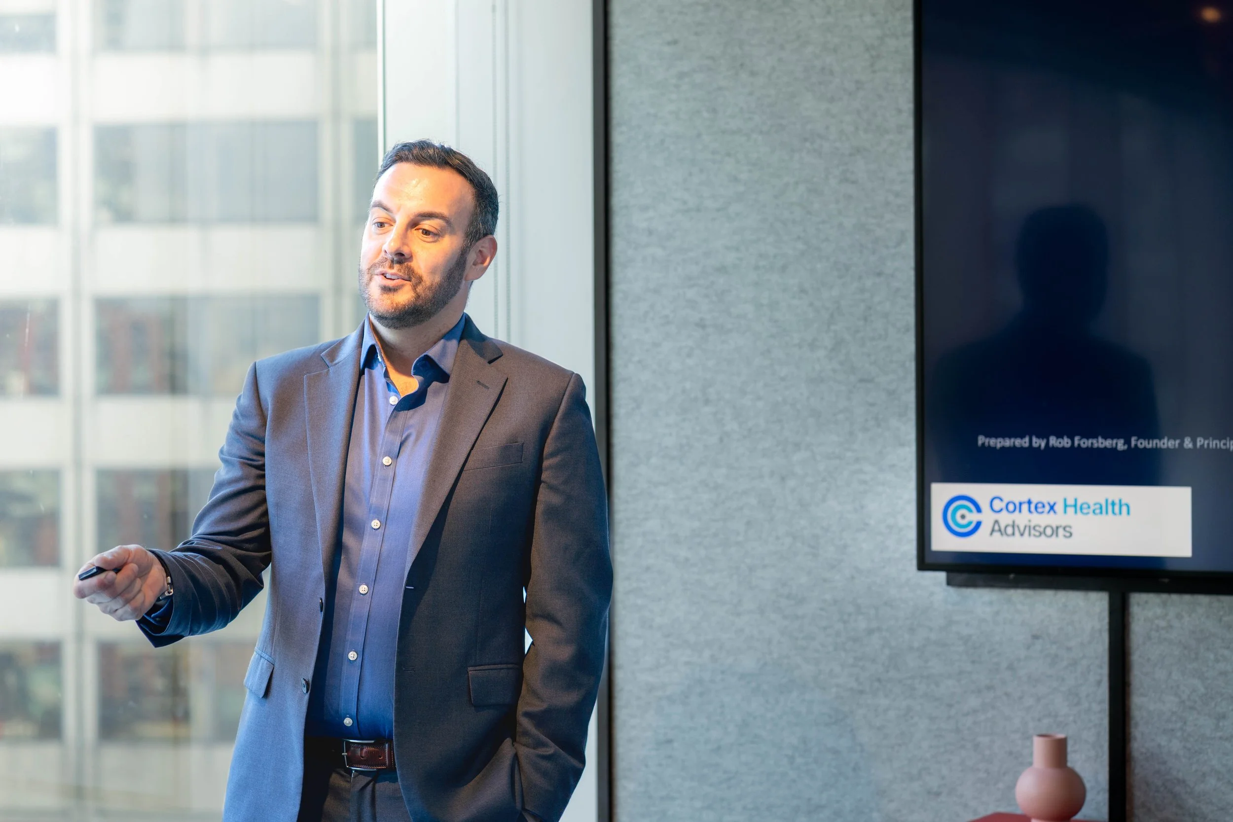 A man in a suit holding a remote control, standing in front of a presentation screen with the Cortex Health Advisors logo, in a modern office with large windows.
