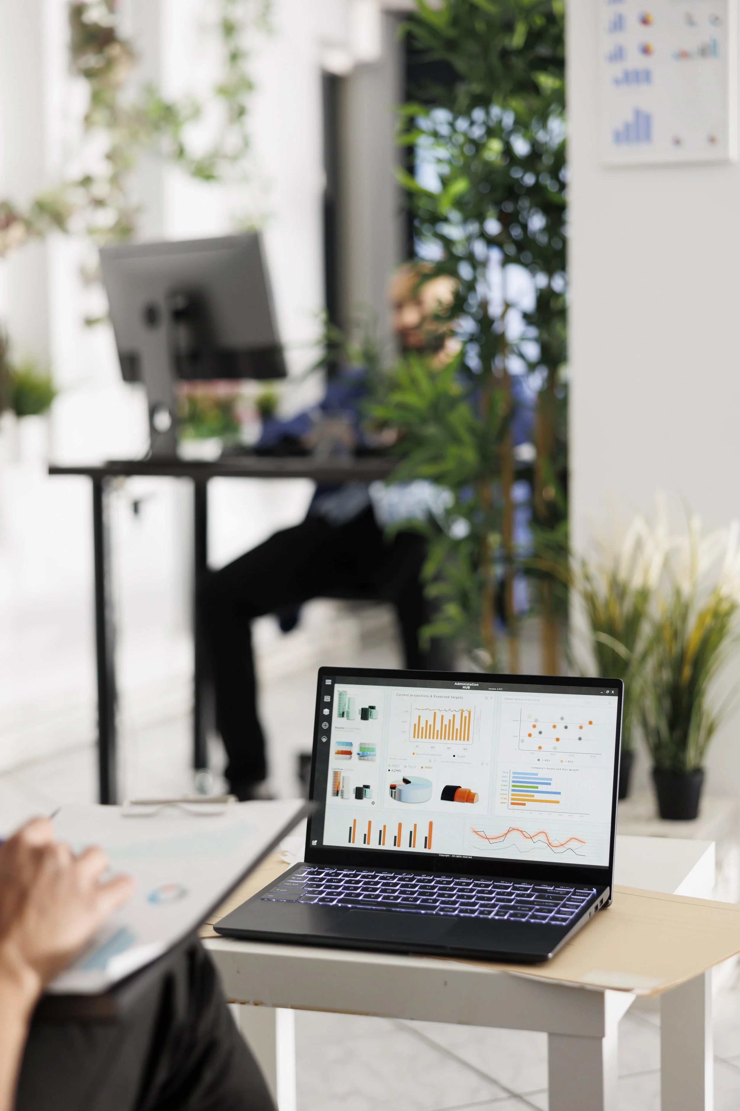 An office setting with a blurred background of a person working at a desk, plants, and charts on the wall. In the foreground, a laptop screen displays various data visualizations and graphs.