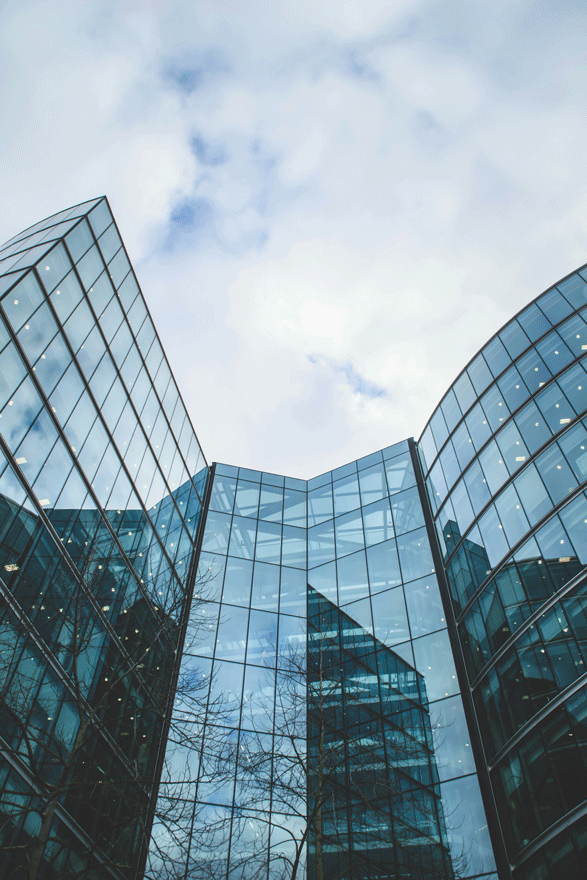 Modern glass office building with curved and angular sections, reflecting the cloudy sky and nearby structures.