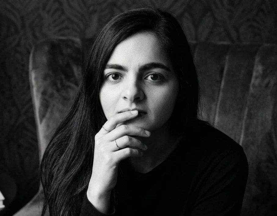 Black and white portrait of a woman with long dark hair, sitting in a chair, looking directly at the camera with her hand near her mouth.