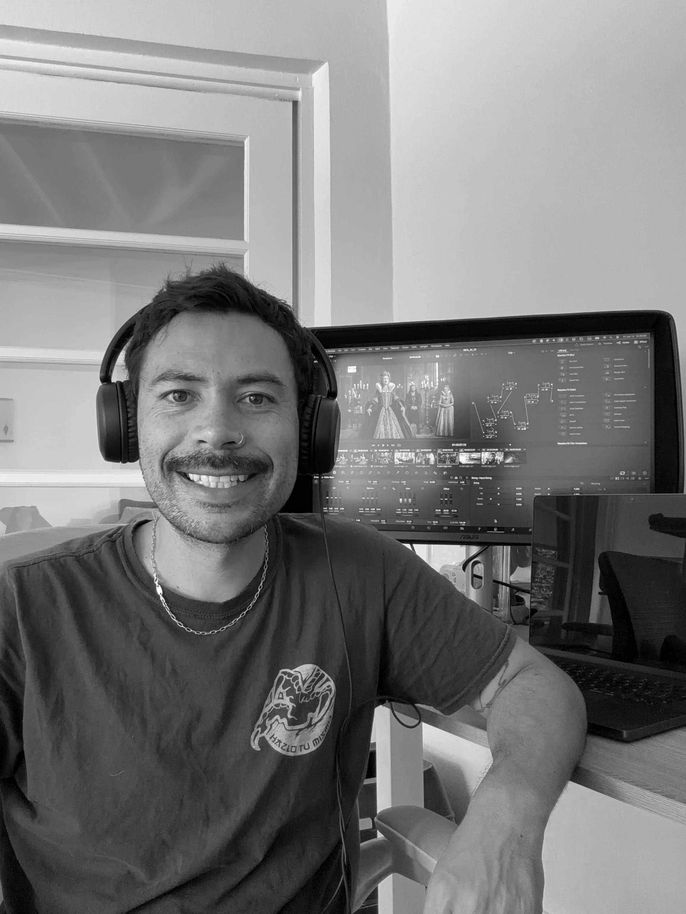 A man with dark hair, wearing headphones, smiling at the camera, seated at a desk with a computer monitor displaying a digital art project or animation and a laptop.