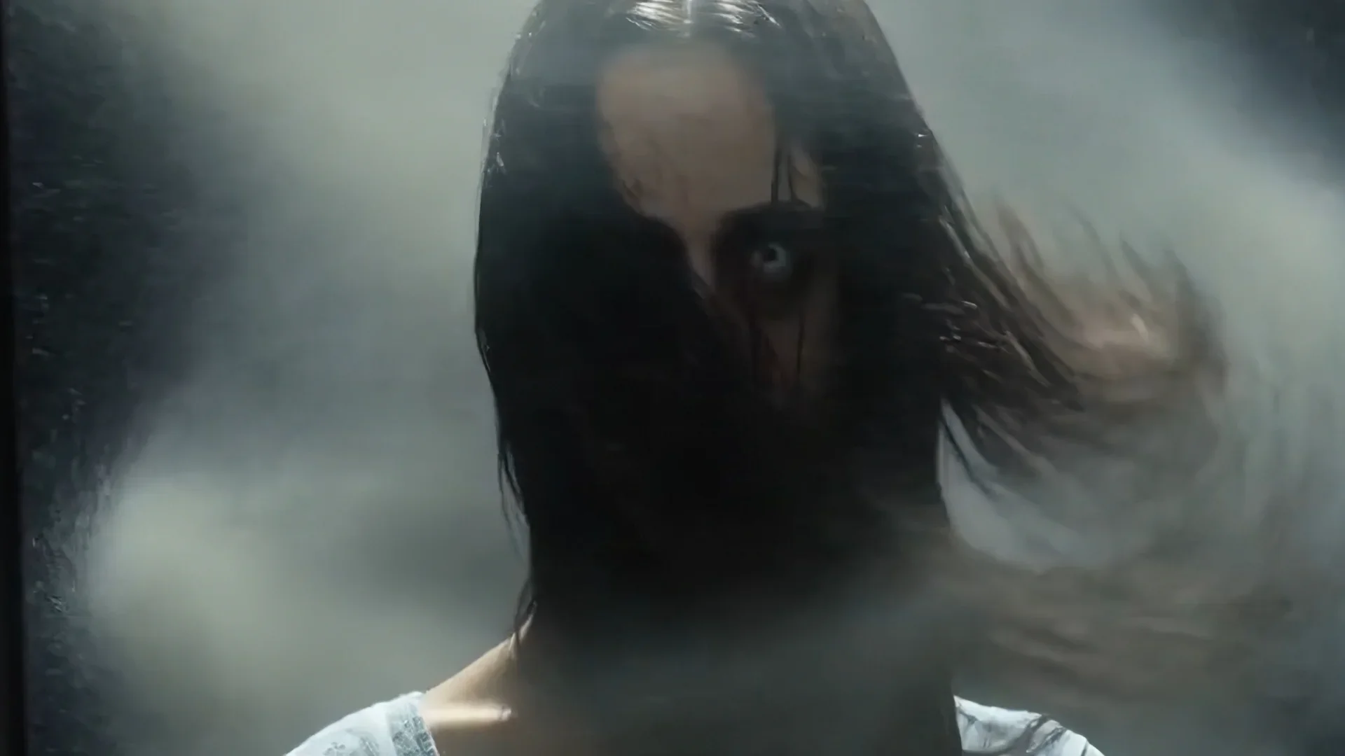 A person with long, wet dark hair and glasses, wearing a white shirt, standing in front of a foggy or misty background.