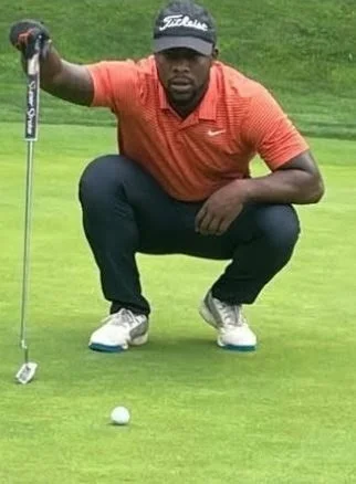 A man crouches on a golf course, holding a golf club, with a golf ball on the green in front of him.