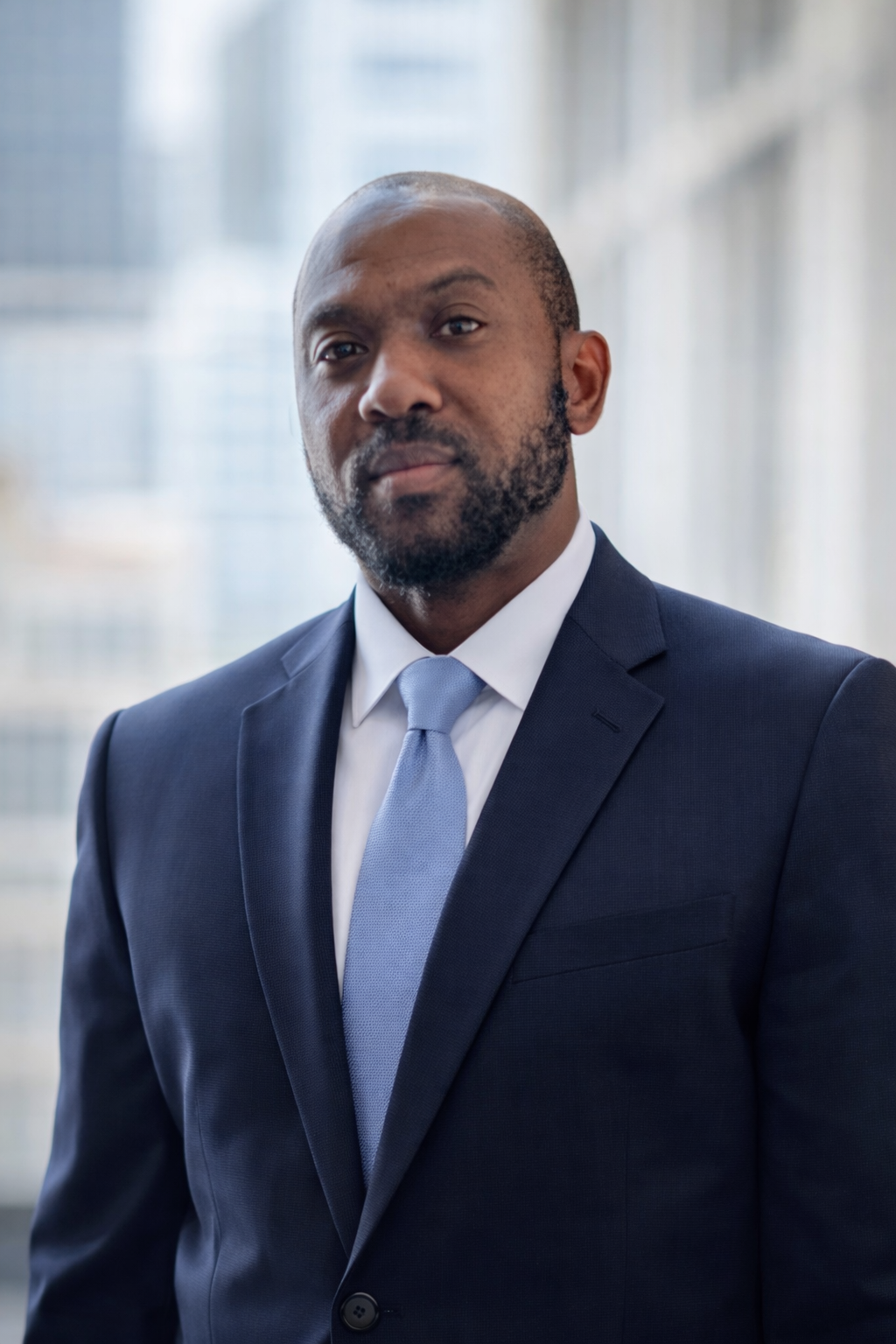 Portrait of a man in a navy blue suit with a light blue tie, standing in front of a window with a blurred cityscape background.