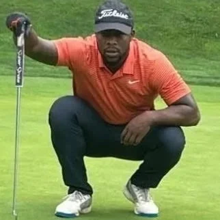 A man crouching on a golf course, holding a golf club with a focused expression.