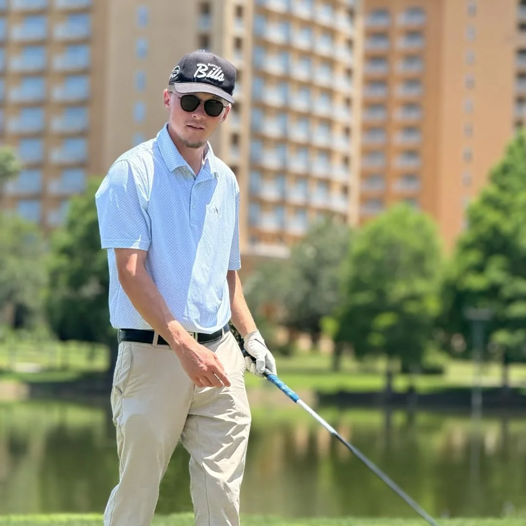 Man playing golf near a pond with tall apartment buildings in the background.