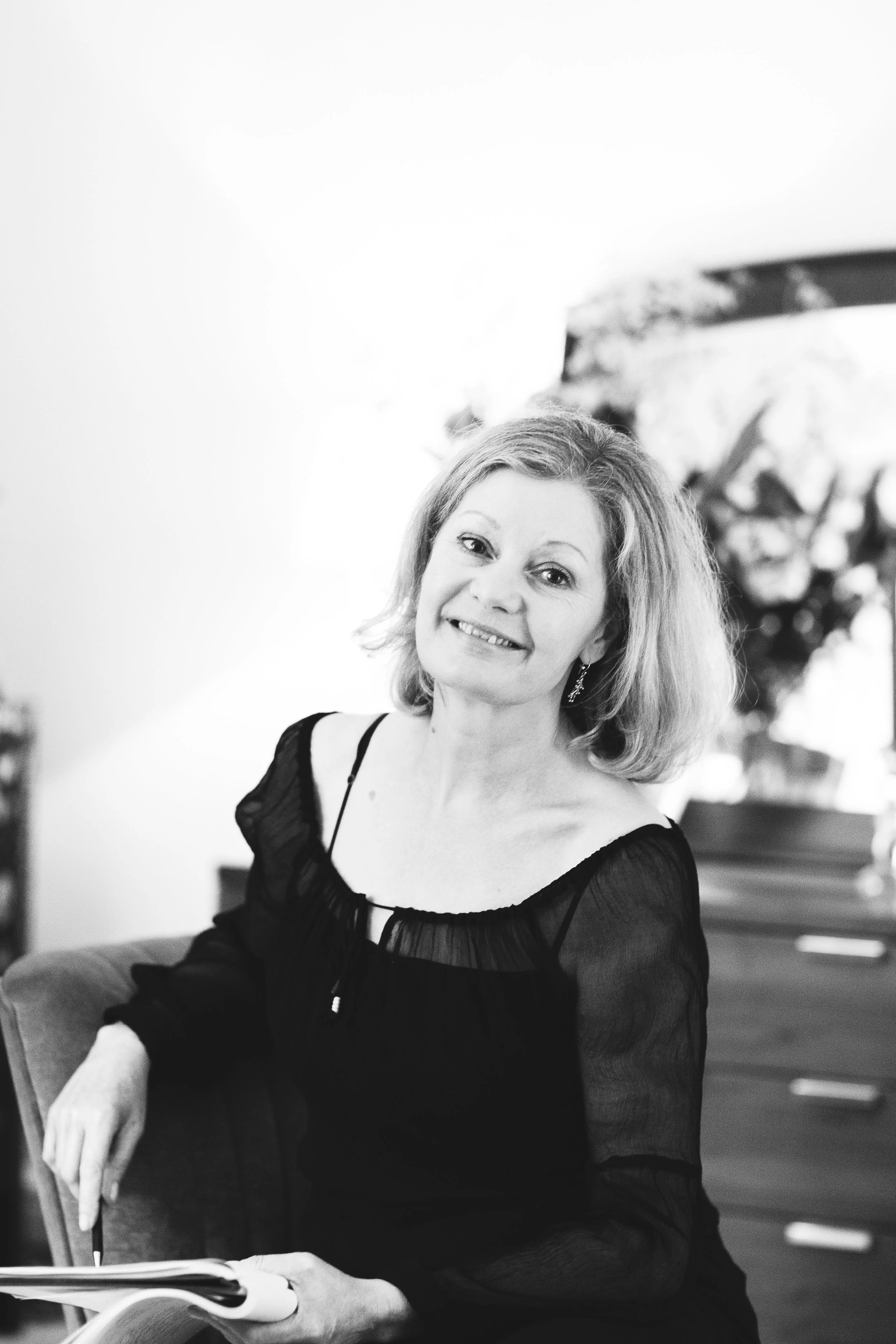 A woman with shoulder-length hair wearing a black dress with sheer sleeves holding a pen and a notebook, sitting on a chair in a room with a painting in the background.