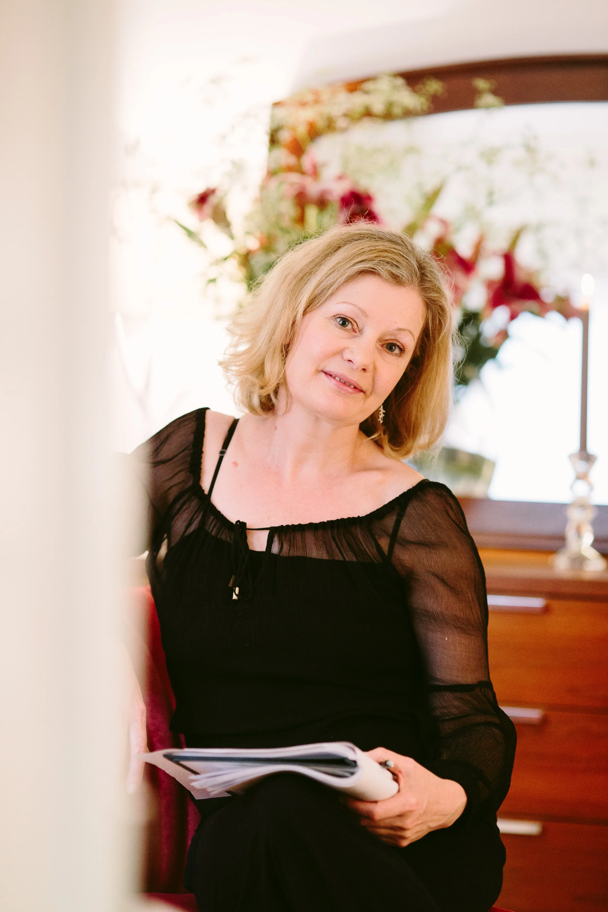 A middle-aged woman with blonde hair wearing a black sheer dress with puffed sleeves, sitting in a room with a floral painting and wooden furniture, holding a book or magazine in her hands.
