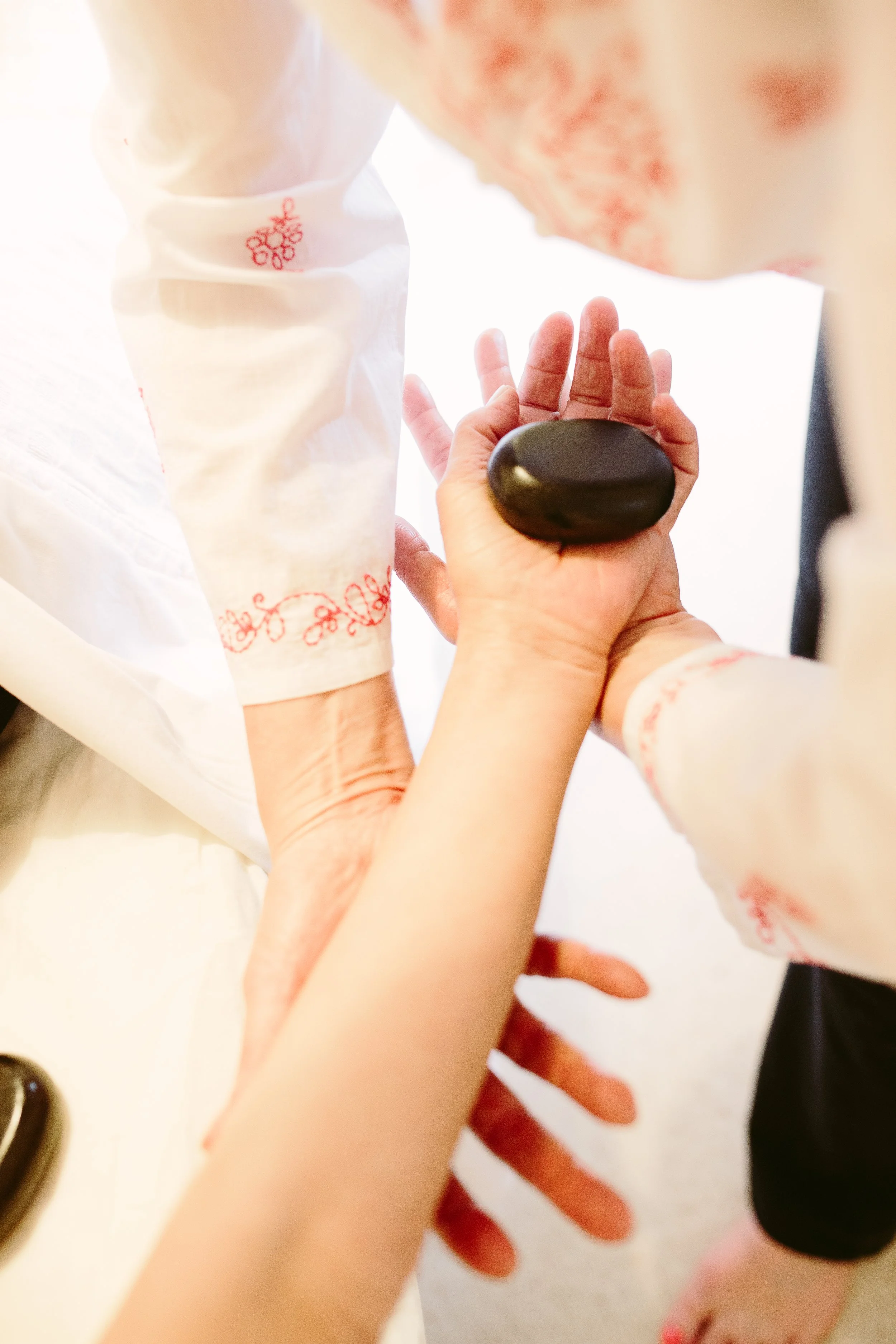 Close-up of a person's hand holding a smooth black massage stone on the palm, with another person's hand gently touching the stone. The background shows part of a person's arm and a soft, white surface.