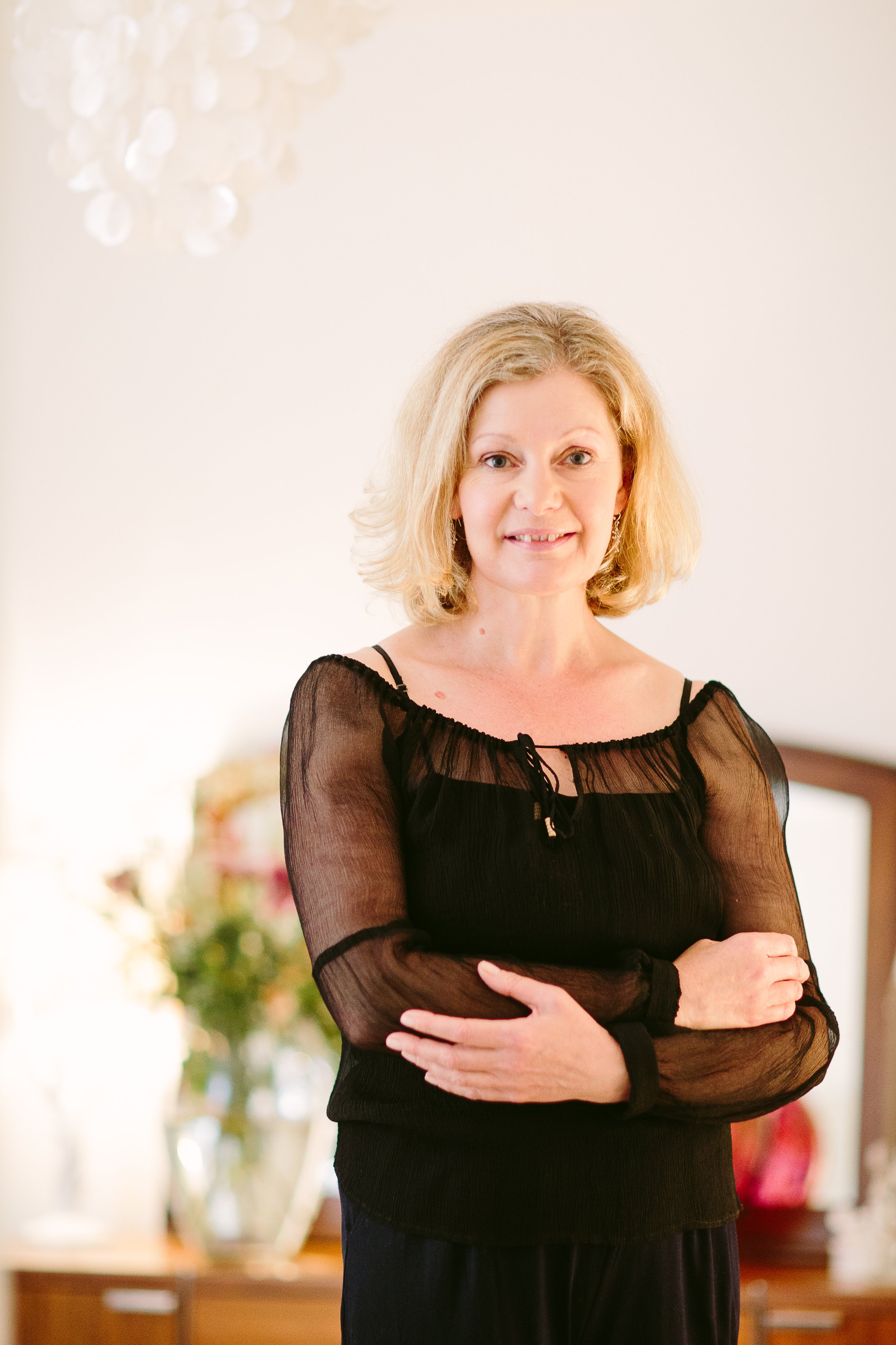 A woman with blonde hair wearing a black sheer top with long sleeves, standing with arms crossed in a room with blurred background including a floral arrangement and a wooden table.