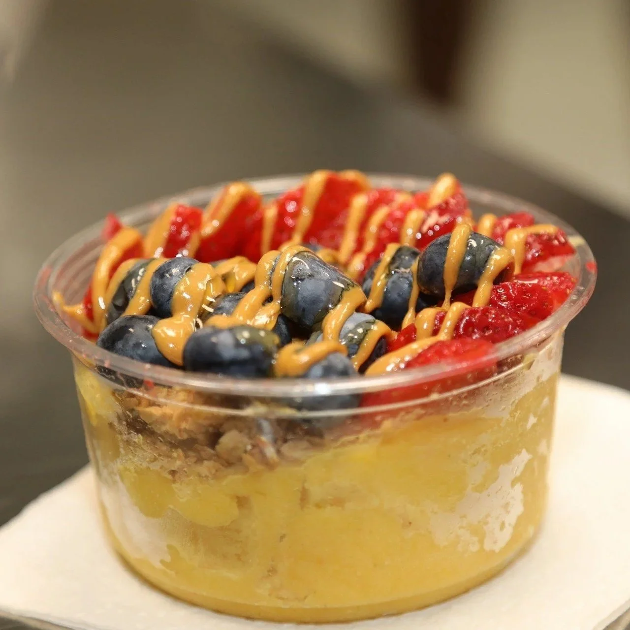 Dessert in a clear plastic cup topped with blueberries, sliced strawberries, and drizzled peanut butter.