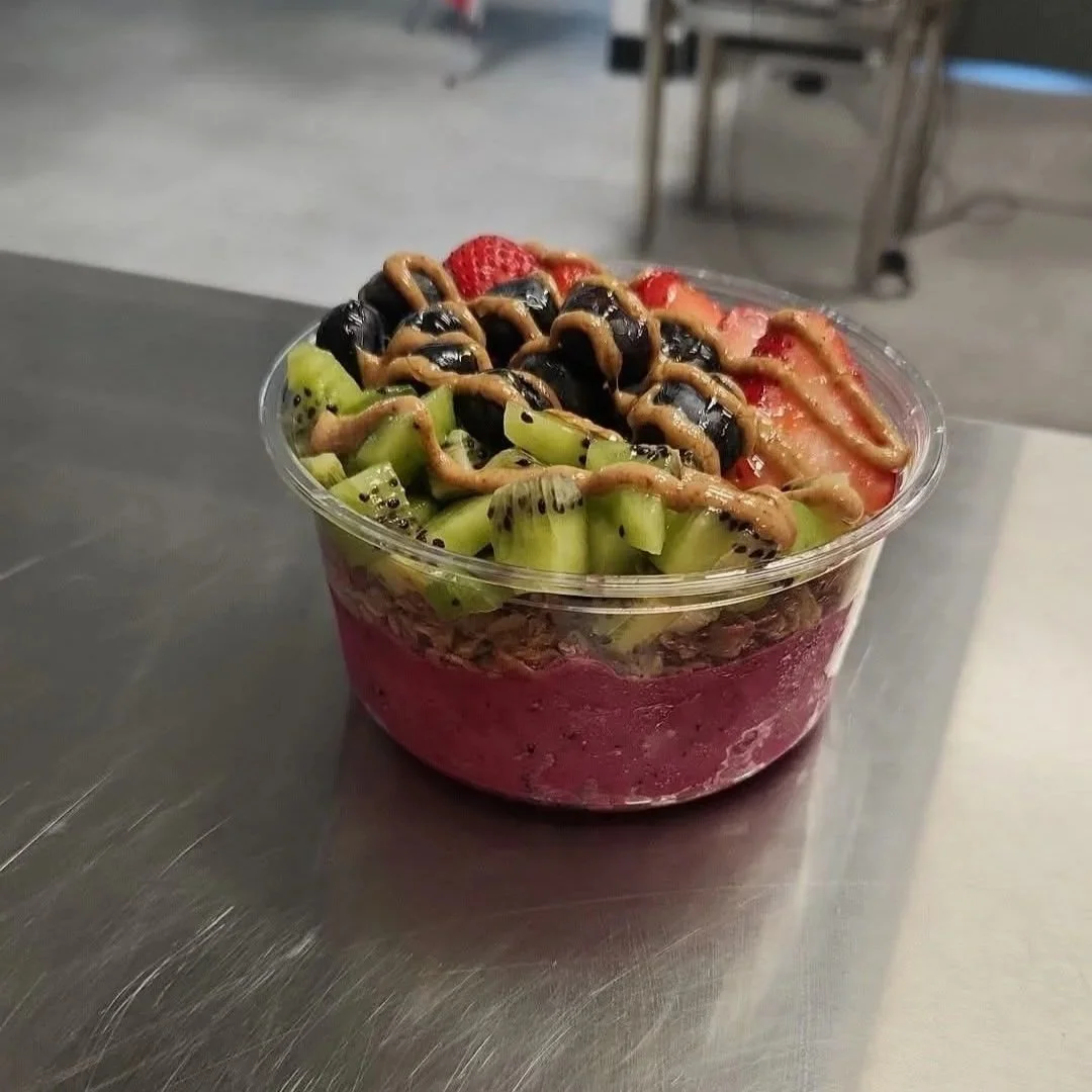 A clear plastic cup filled with layered fruit, topped with strawberries, blueberries, kiwi, and drizzled with chocolate and peanut butter sauces.