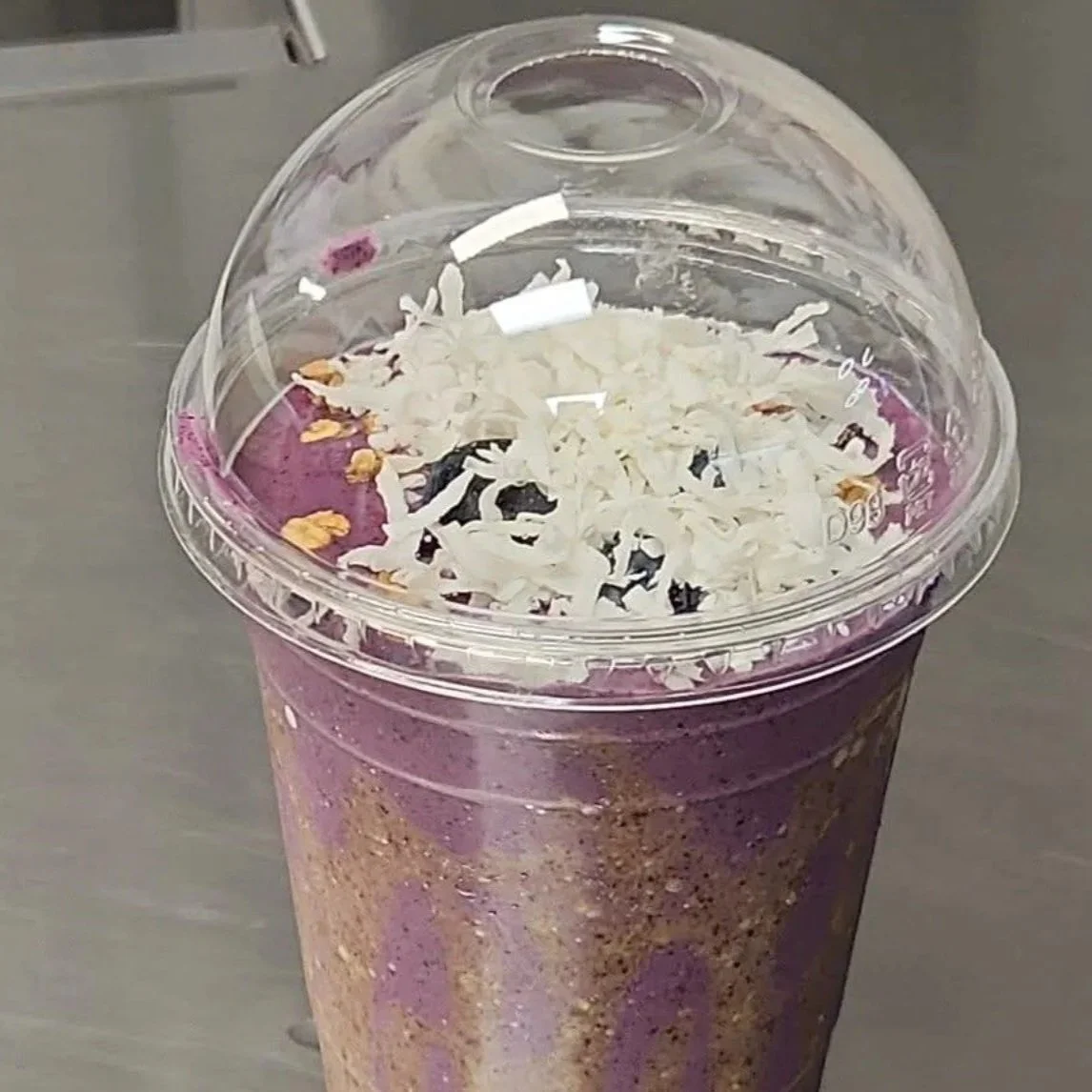 A purple blended smoothie in a clear plastic cup topped with shredded white cheese under a clear plastic dome lid.