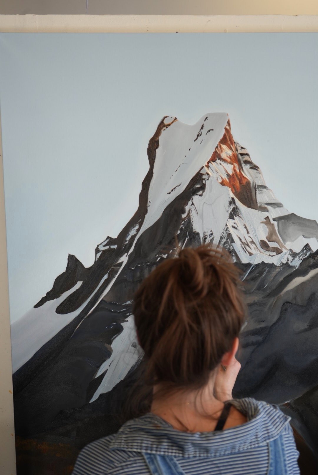 A woman with short brown hair wearing a striped shirt and denim apron is looking at a painting of a snow-capped mountain.
