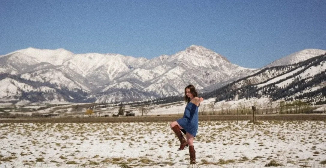 A woman in a blue dress and brown boots standing in a snowy field with mountains in the background.