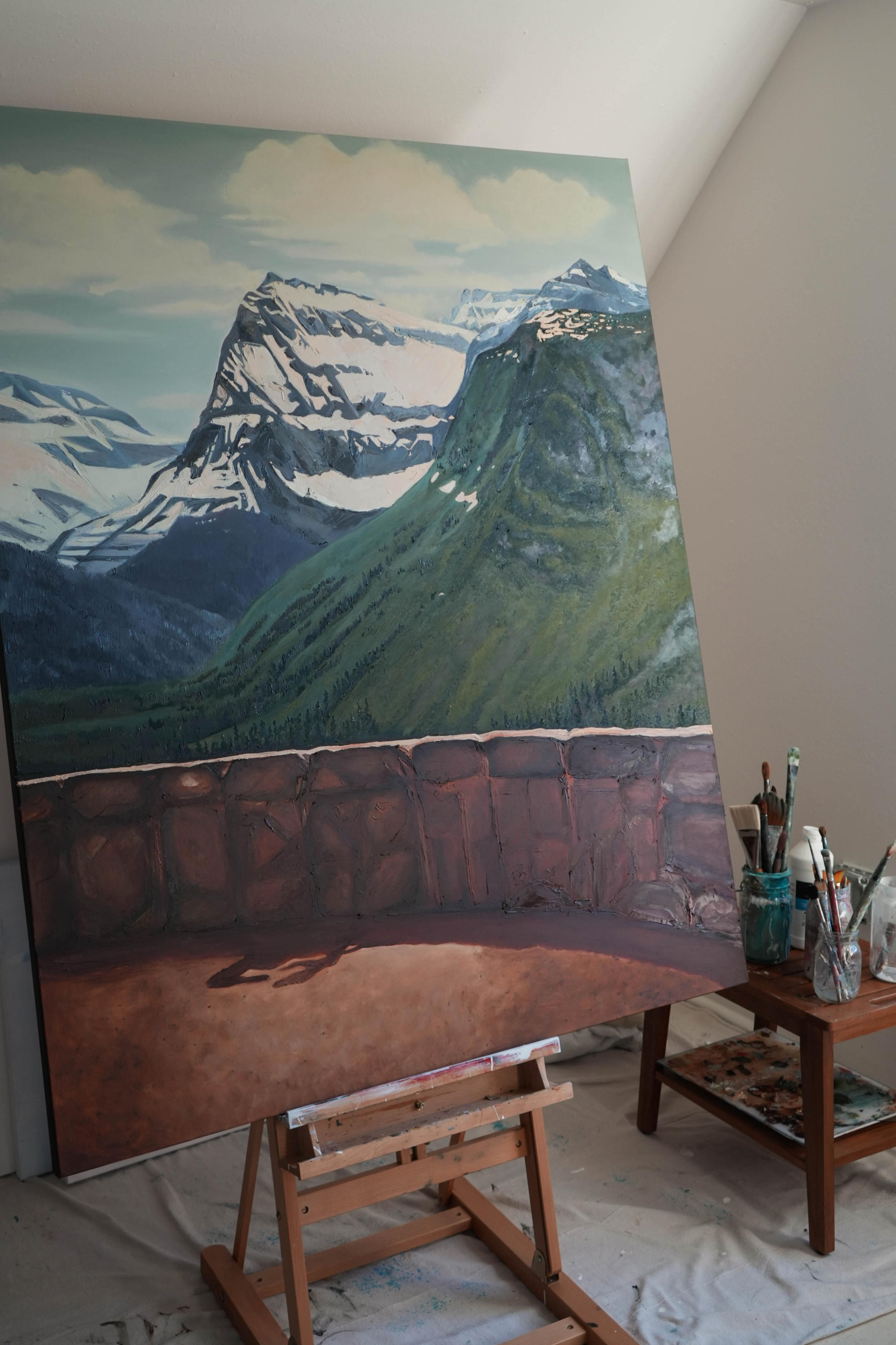 A painting of a mountain landscape with snow-capped peaks and green slopes, set in an artist's studio with paints and brushes nearby.
