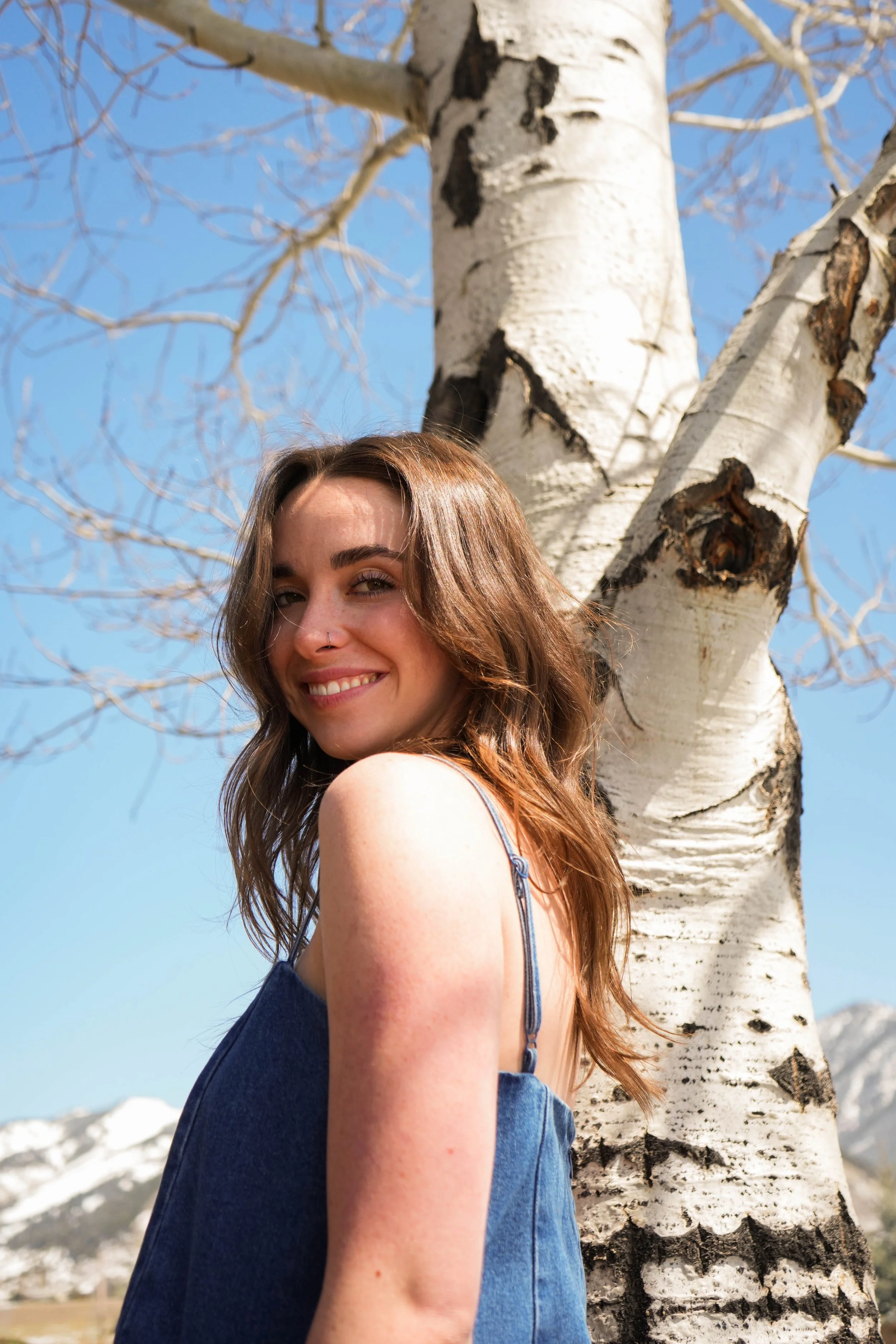 Young woman with long brown hair smiling while standing next to a white birch tree in a mountainous outdoor setting with snow-covered peaks in the background.