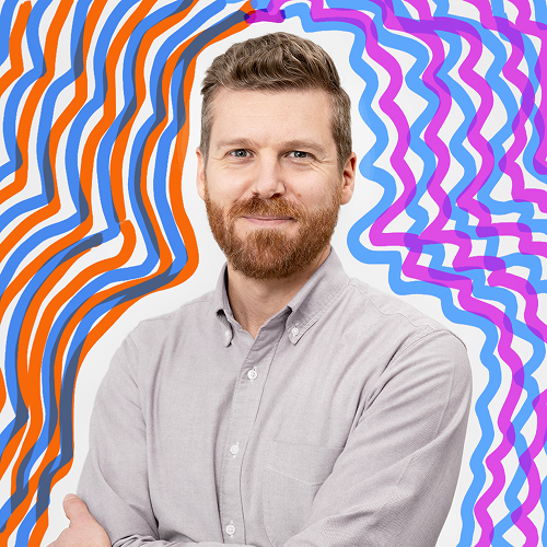 Portrait of a man with a beard and short hair, wearing a light gray shirt, standing in front of a colorful, abstract, wavy patterned background.